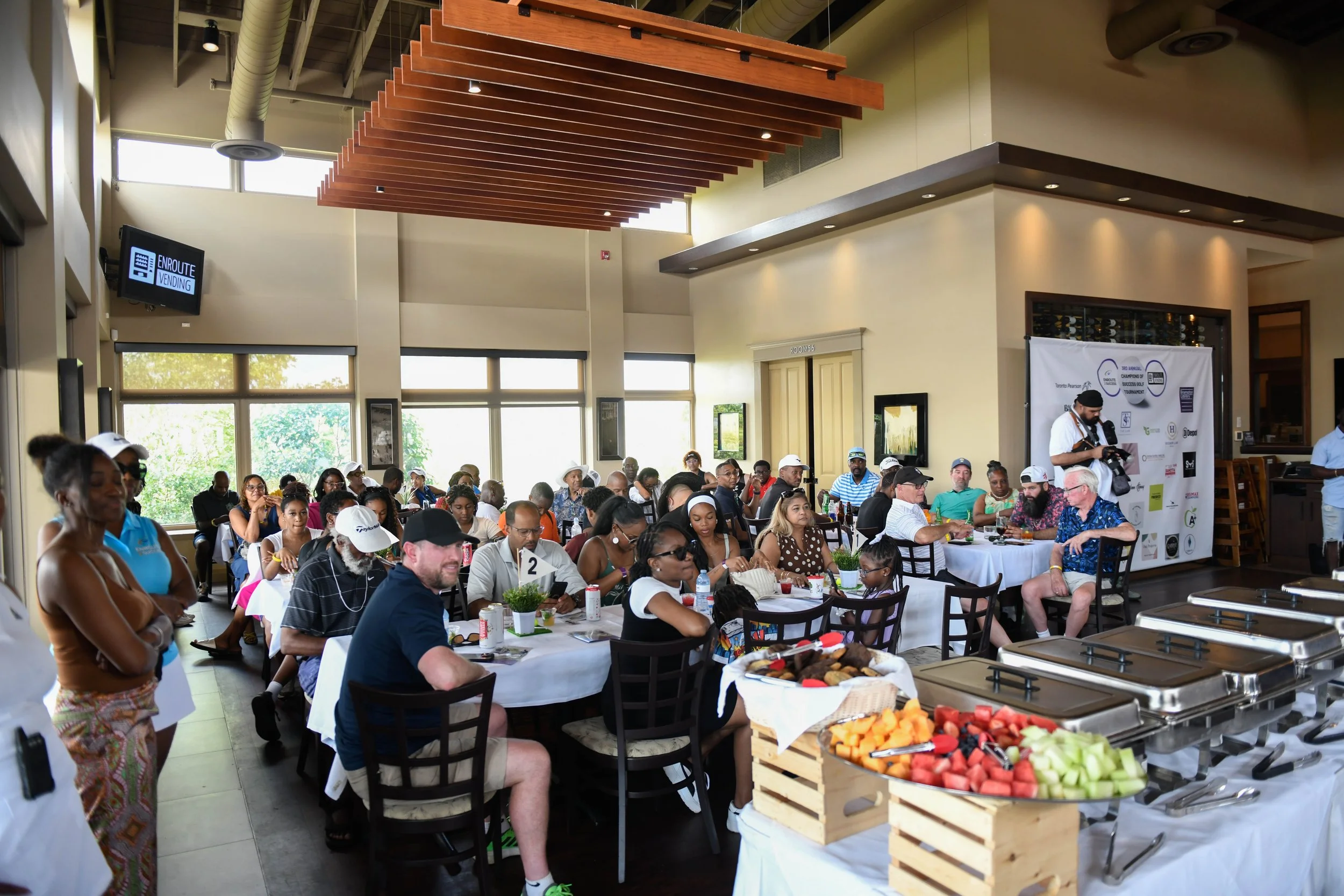 Attendees sit together during an Enroute 2 Success golf tournament, gathered around tables with food and refreshments while listening to a presentation in a bright indoor venue.