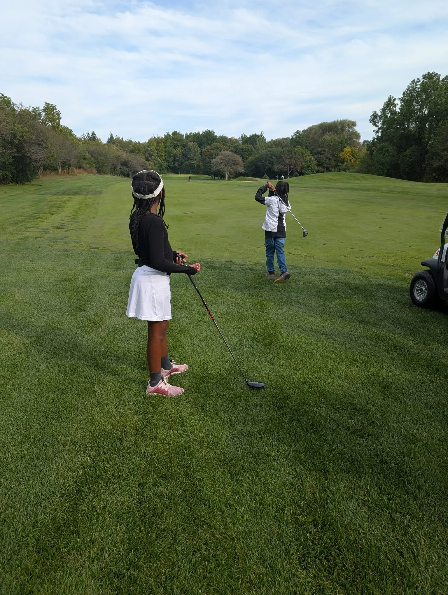 Two young golfers participating in EnRoute 2 Success youth golf activities, with one practicing her swing on a green fairway while the other watches with her golf club.