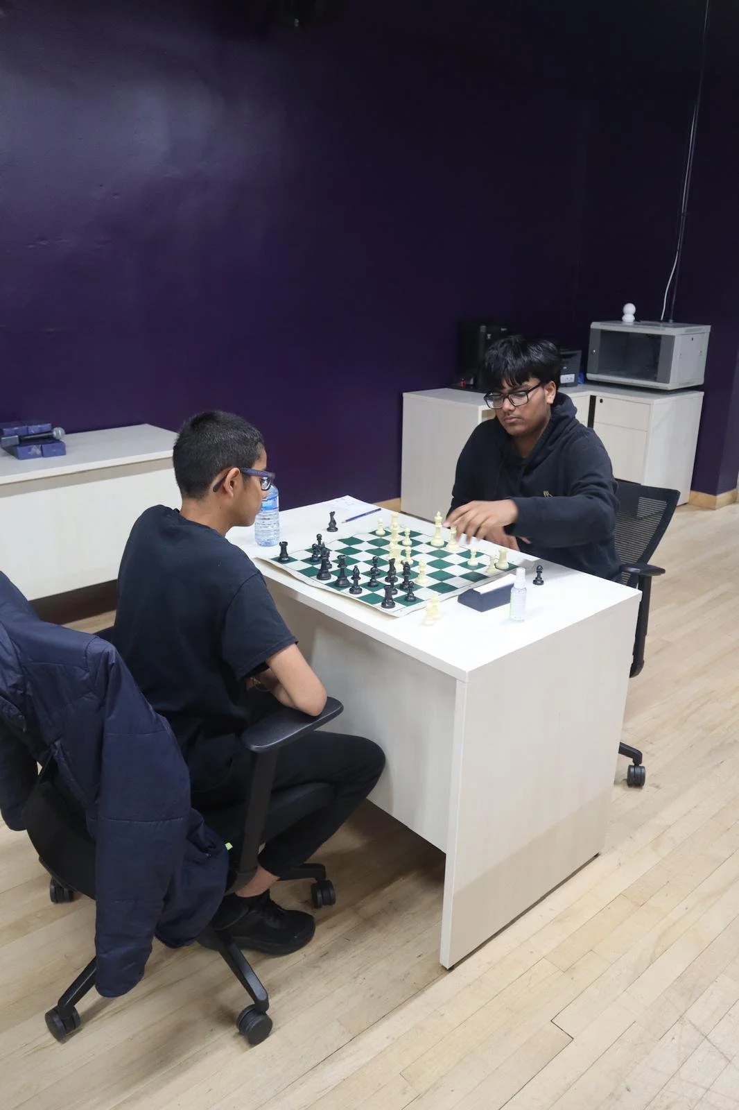 Two youth participants sit across from each other at a desk playing chess during an EnRoute 2 Success session, focused on the board in a room with purple walls and classroom furniture.