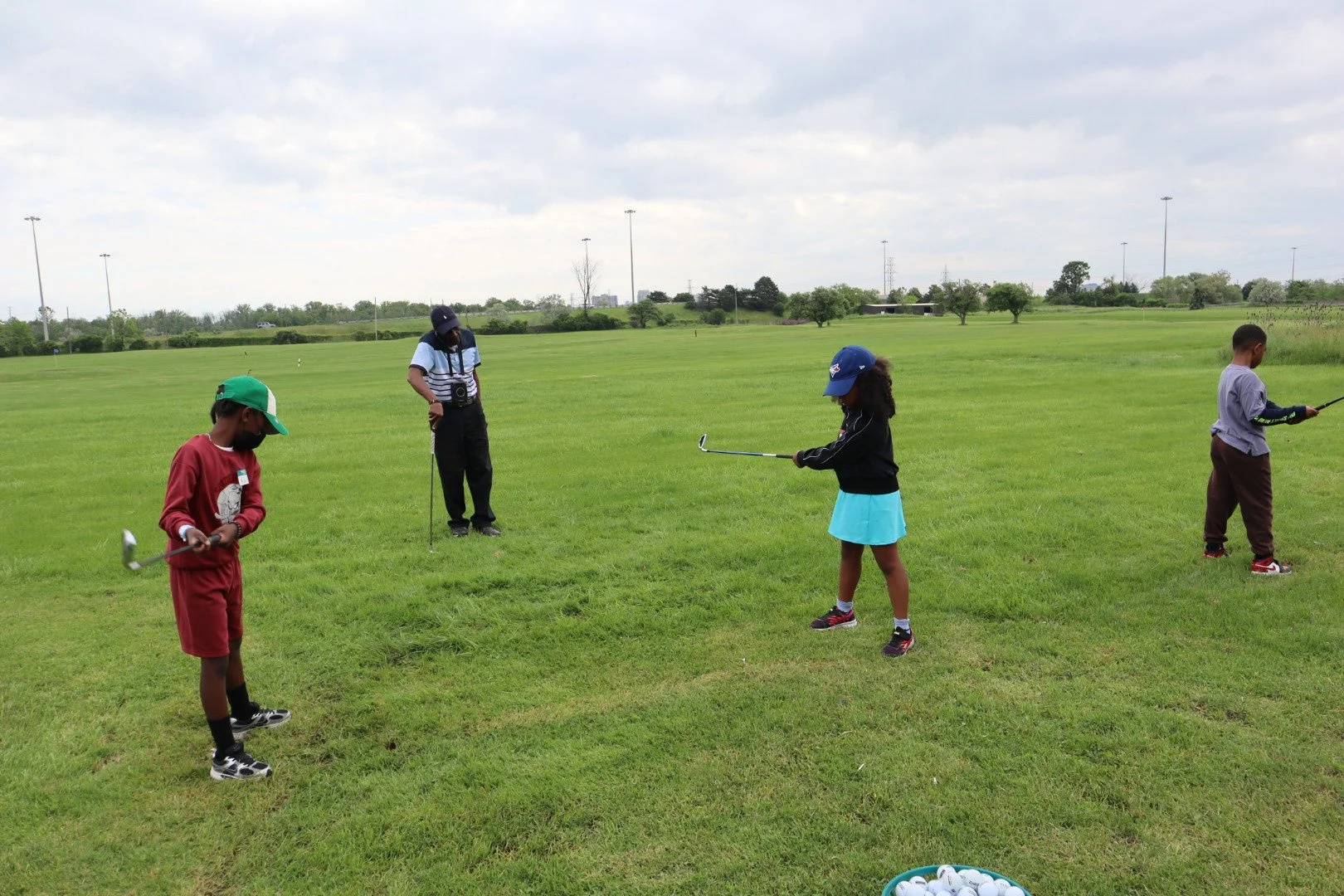 Three youth practice their golf swings on a grassy driving range during an EnRoute 2 Success golf lesson. An instructor stands nearby observing, with a basket of golf balls in the foreground.