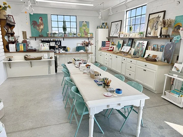 Art studio with a long white table surrounded by teal chairs, artwork on walls, and art supplies on the table and shelves.