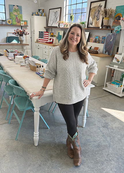 A woman smiling and posing in an art studio with paintings on the walls, a long table with art supplies, and art displayed on shelves.