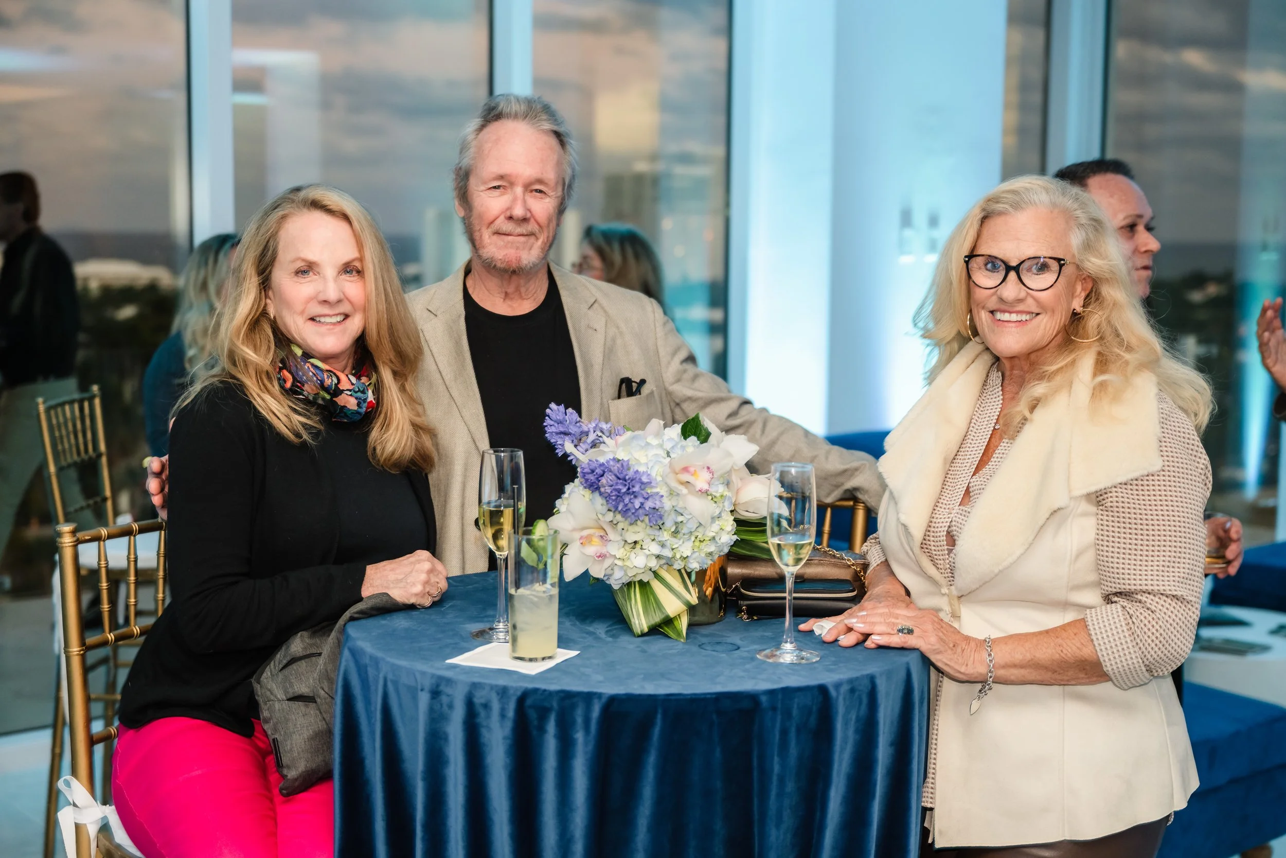 Four adults, two women and one man, standing around a table with a bouquet of flowers, at a social event in a modern room with large windows showing a cityscape at sunset. They are smiling and holding champagne glasses.