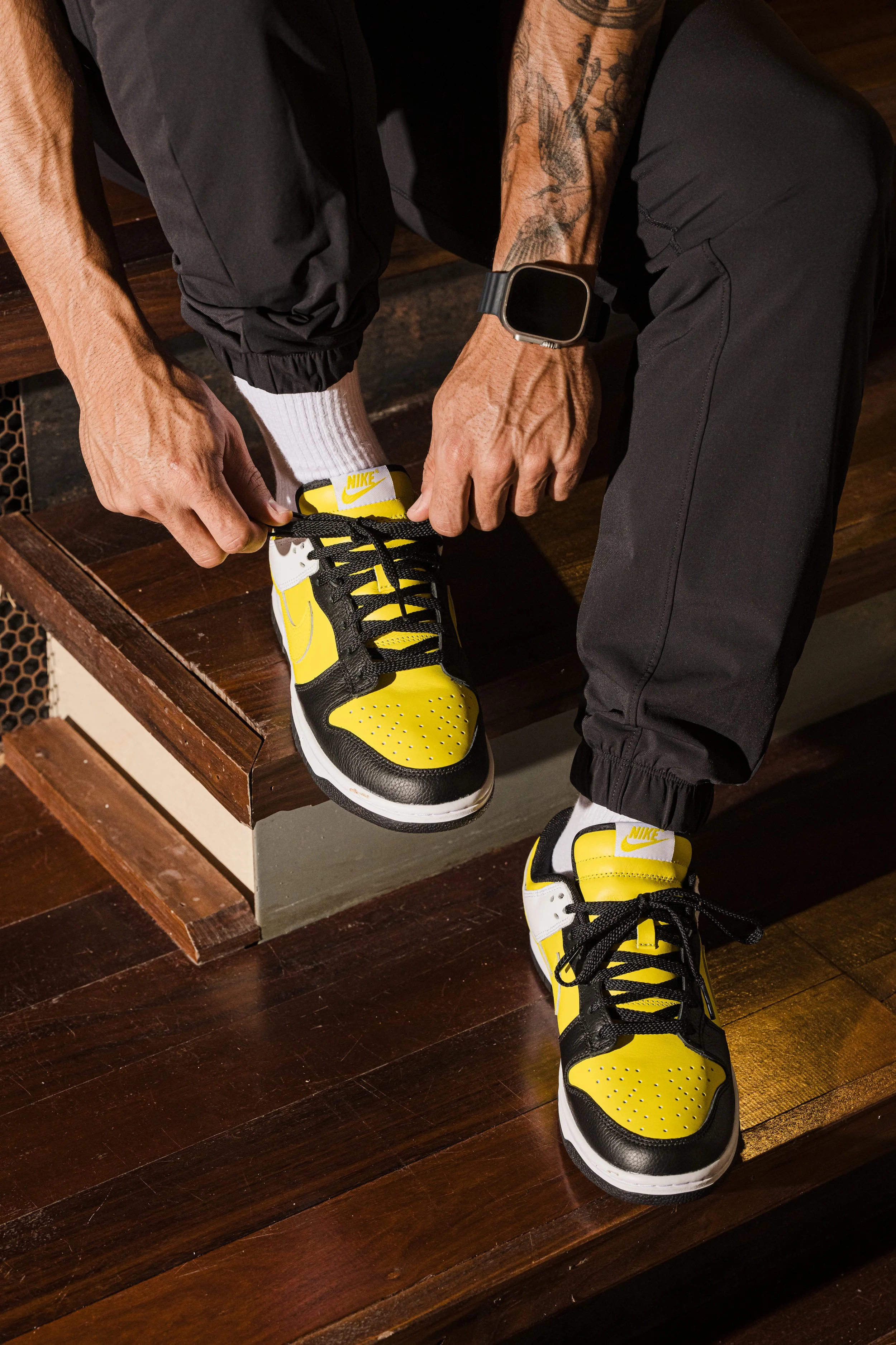A person wearing black pants, a black watch, and white socks is tying the laces on a pair of yellow, black, and white sneakers, while sitting on a wooden bench.