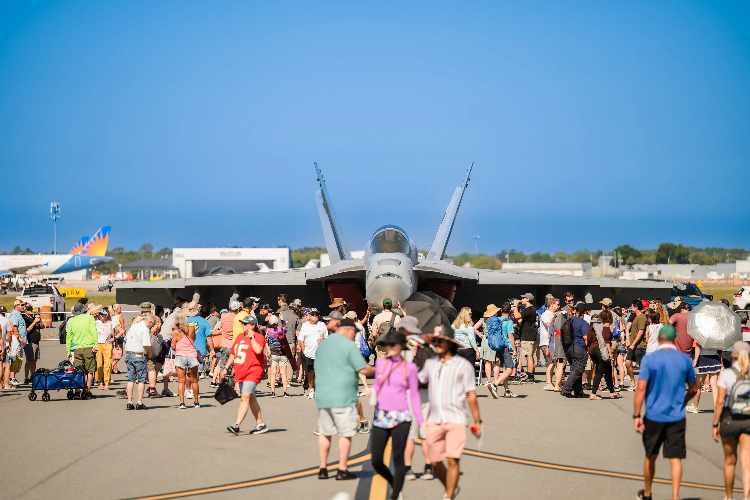 A large crowd of people gathered around a military jet on display at an airshow. The jet is facing directly toward the camera, with other aircraft visible in the background on a clear, sunny day.