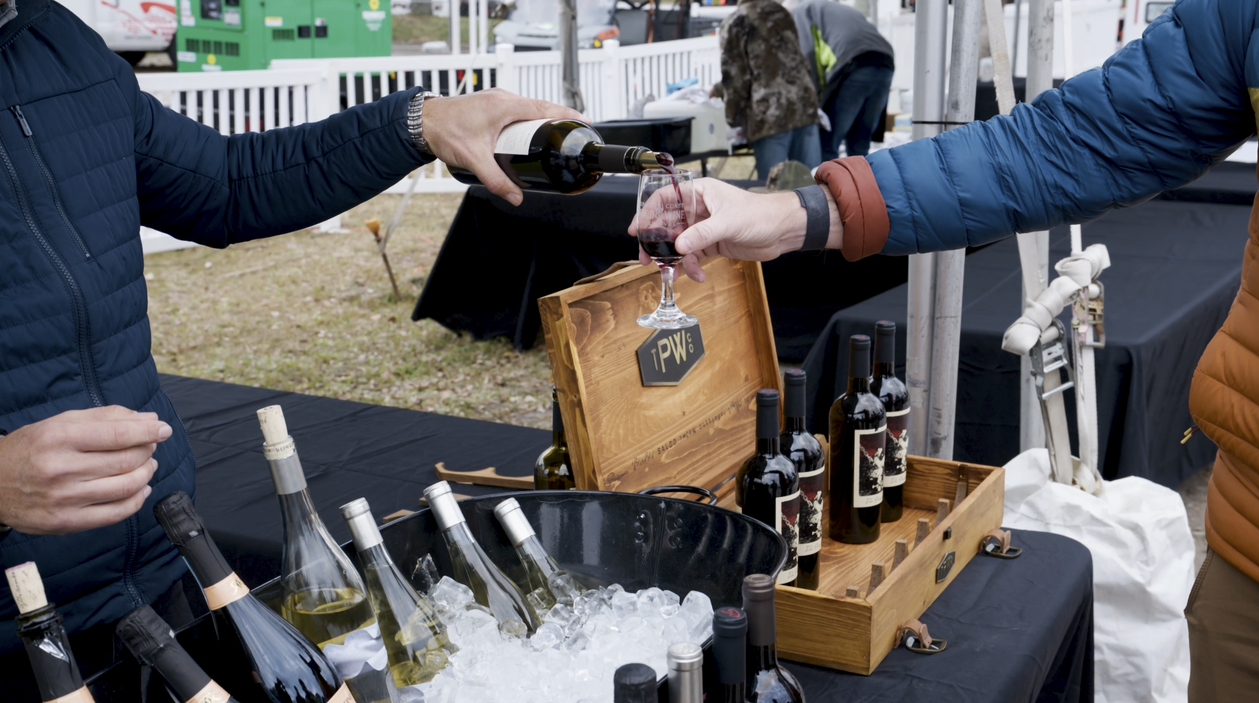 Close-up of a wine vendor pouring red wine into a glass for a festival attendee at an outdoor tasting booth with bottles on ice and a wooden wine crate display at the Space Coast Food & Wine Festival in Melbourne, Florida