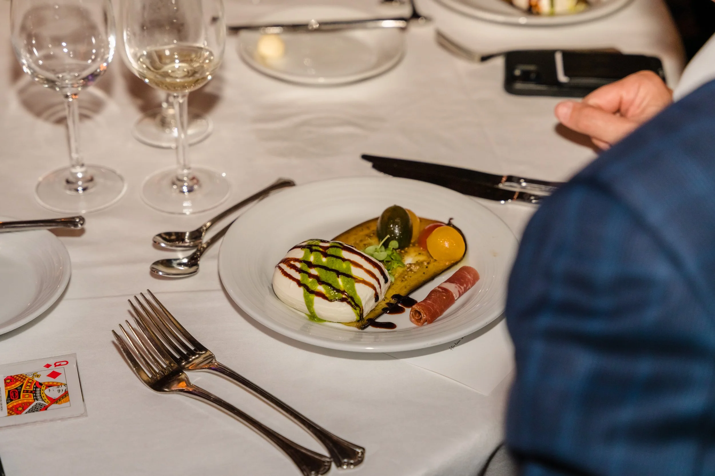 Close-up of an elegantly plated burrata appetizer with pesto drizzle, balsamic reduction, heirloom tomatoes, and prosciutto on a white-clothed table, with a guest's hands clasped beside the plate and wine glasses in the background.