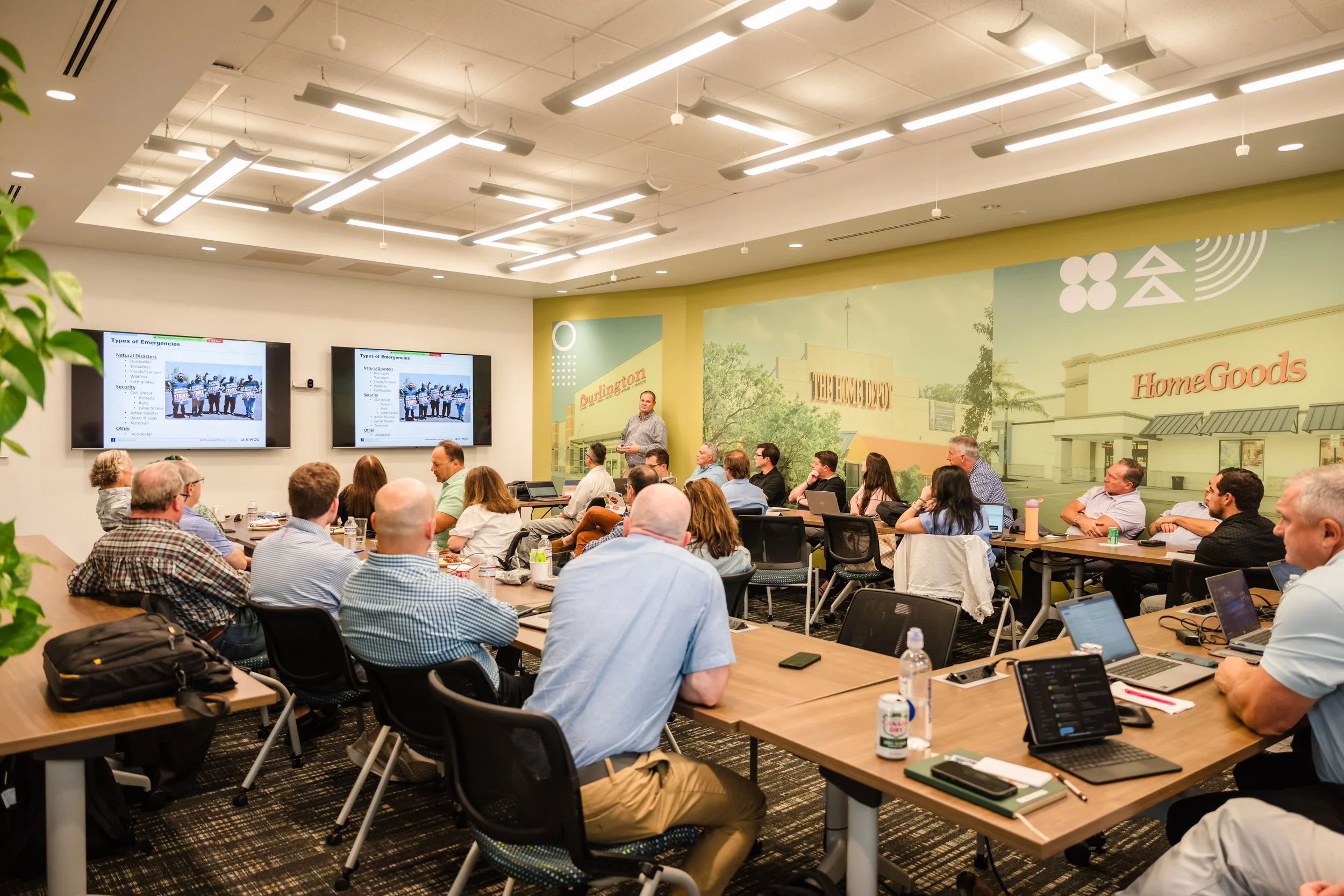 A group of people attending a presentation in a conference room with large digital screens on the wall, decorated with a mural of a shopping mall exterior with signs for HomeGoods and The Home Depot.