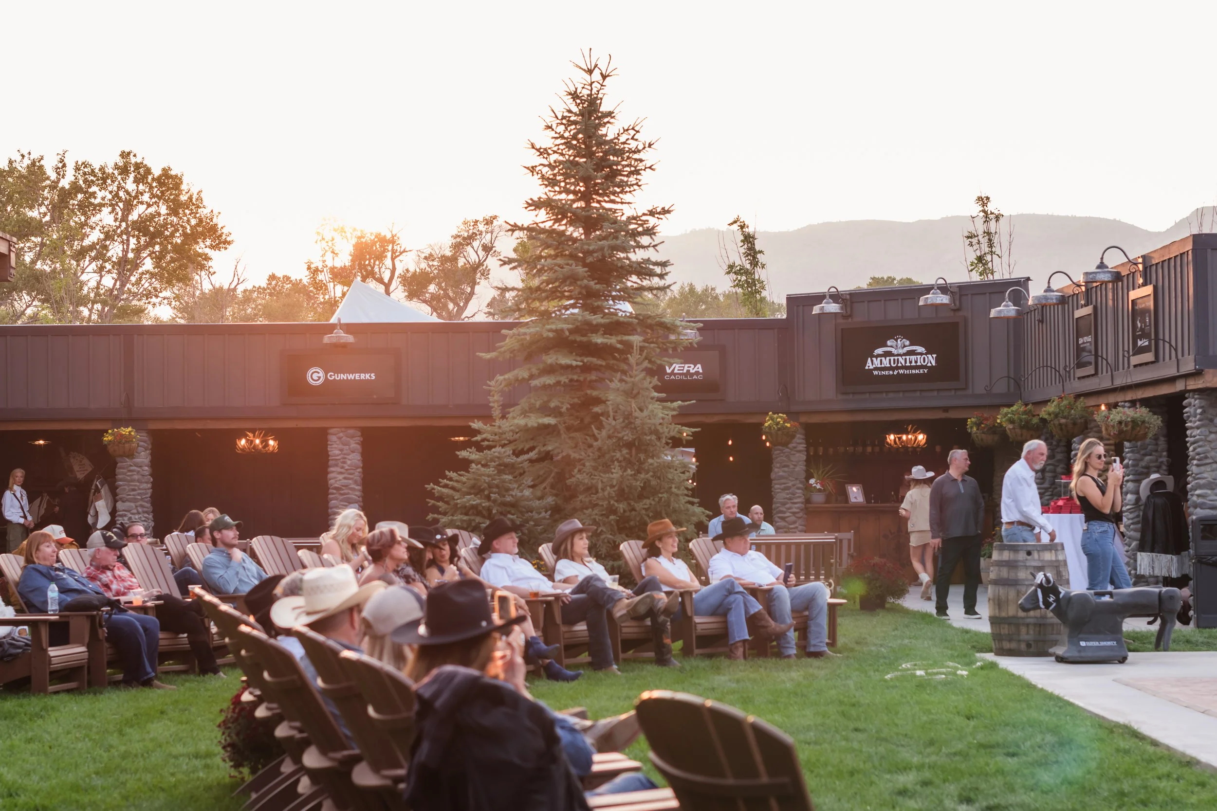 People attending an outdoor event at sunset with a rustic bar in the background and mountains in the distance.