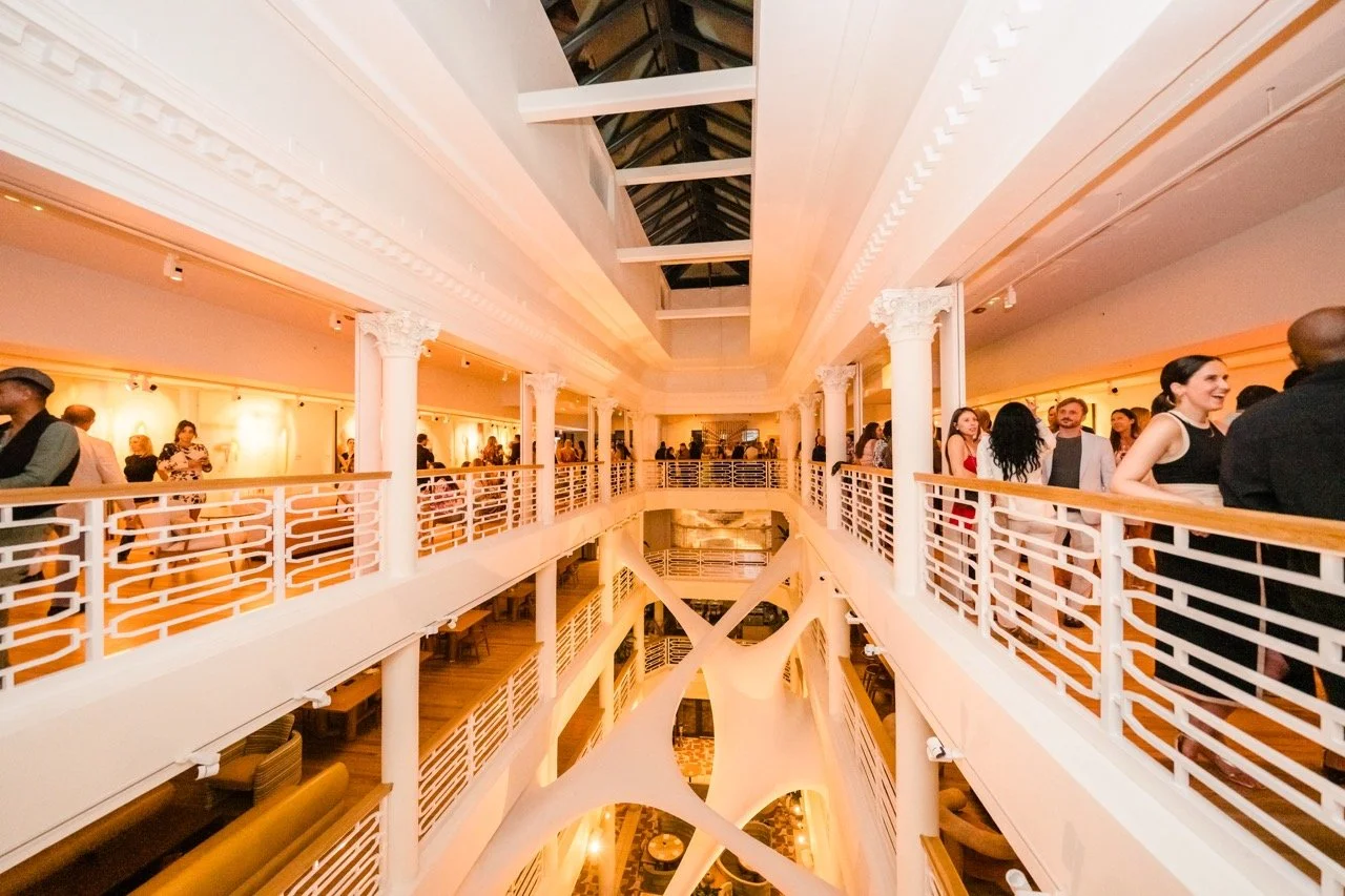 Interior of a multi-level event space with white railings, columns, and a high ceiling, filled with people socializing.