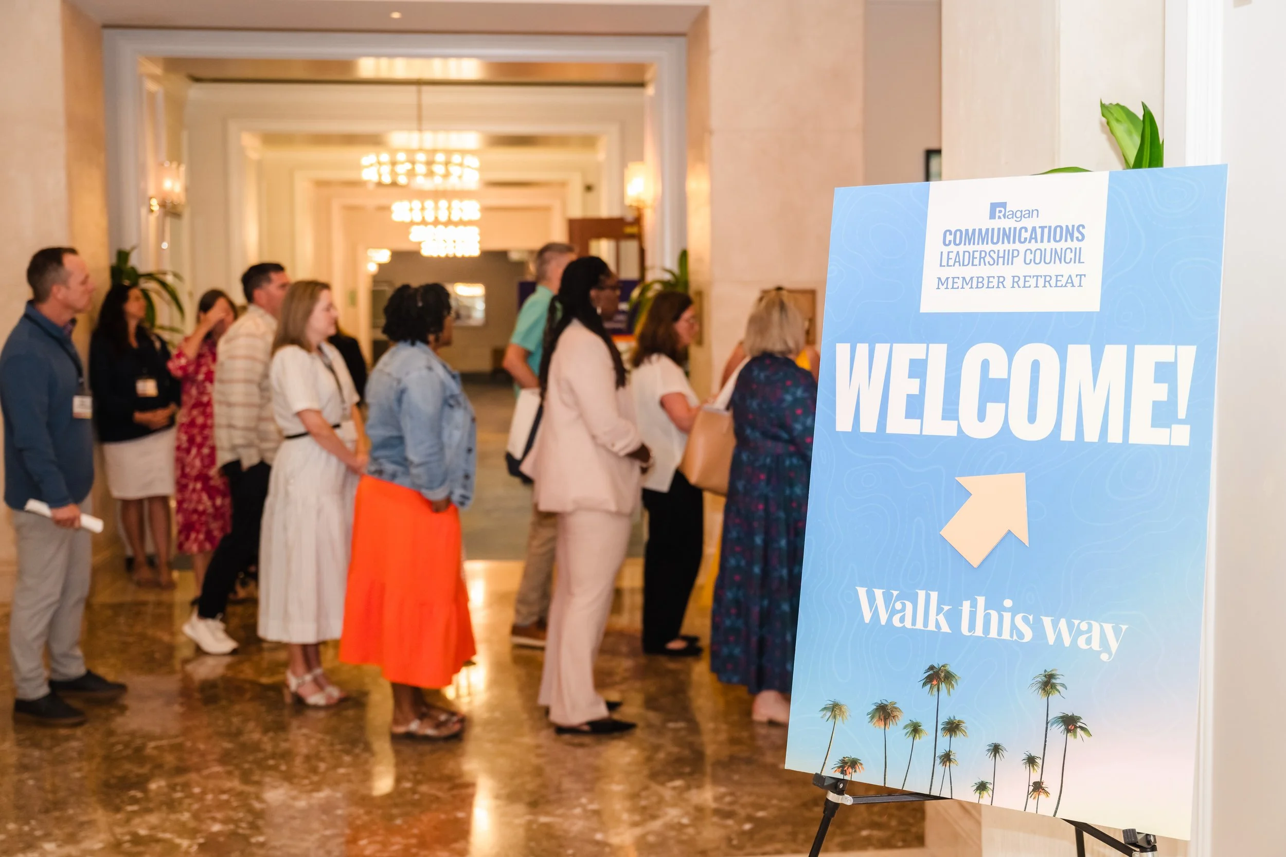 A group of professionals mingles in an elegant hotel lobby beside a blue welcome sign for the Ragan Communications Leadership Council Member Retreat, featuring palm tree graphics and a directional arrow reading "Walk this way."