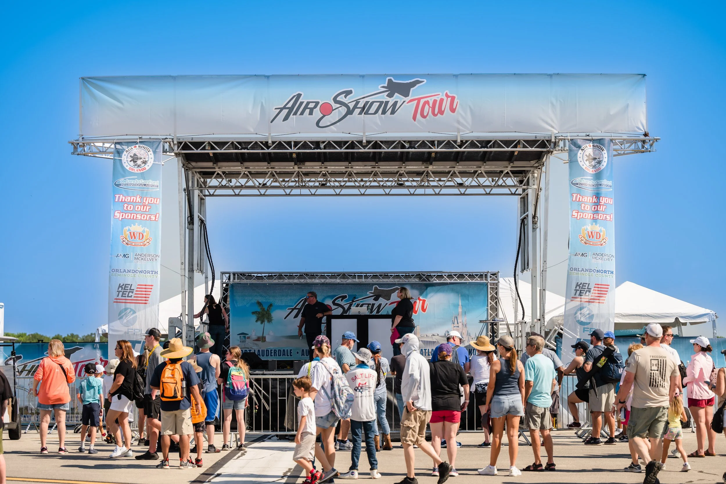 Crowd of people attending an outdoor air show event with a stage and large banners displaying aircraft and sponsor logos under a blue sky.
