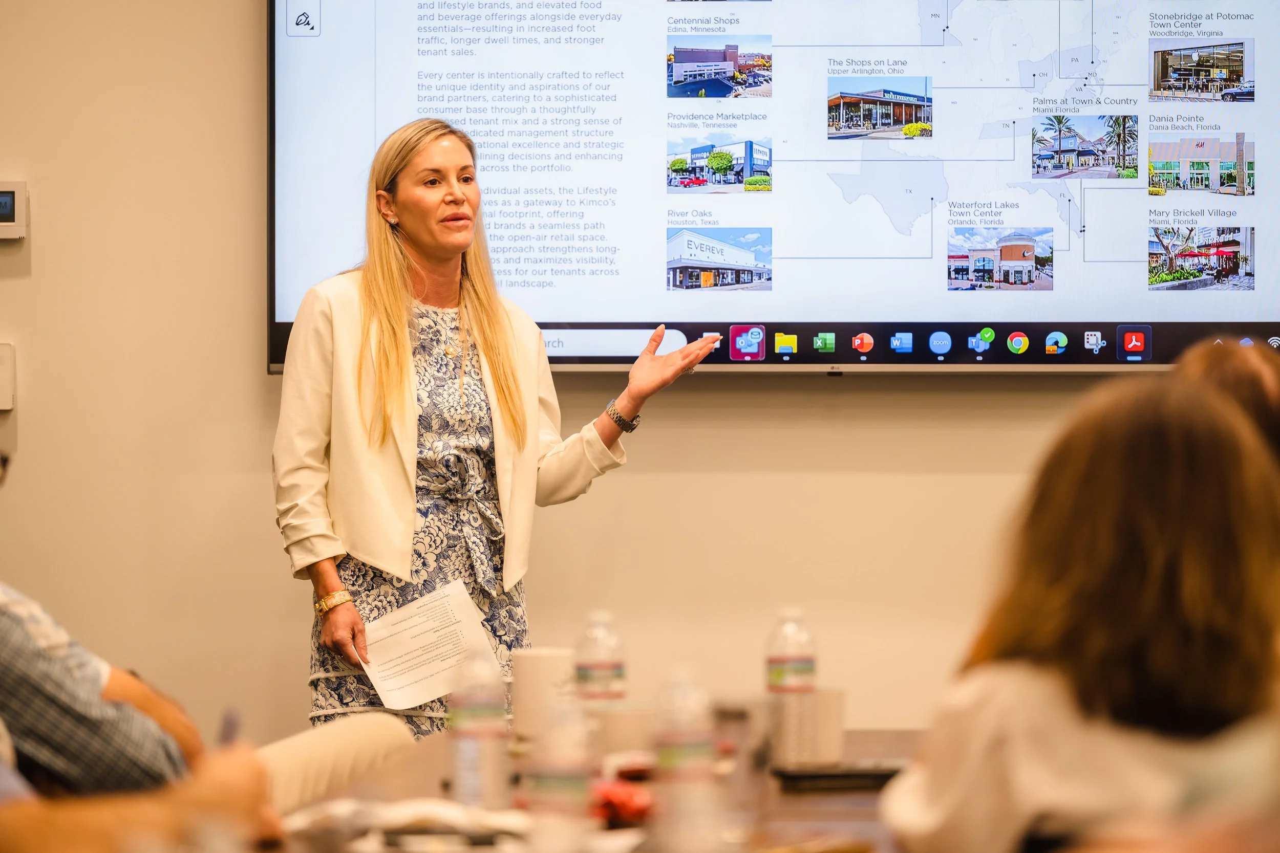 A woman is giving a presentation in a conference room with a large screen displaying buildings and location information behind her. She is holding papers and gesturing with her right hand to the screen, while several attendees are seated and listenin