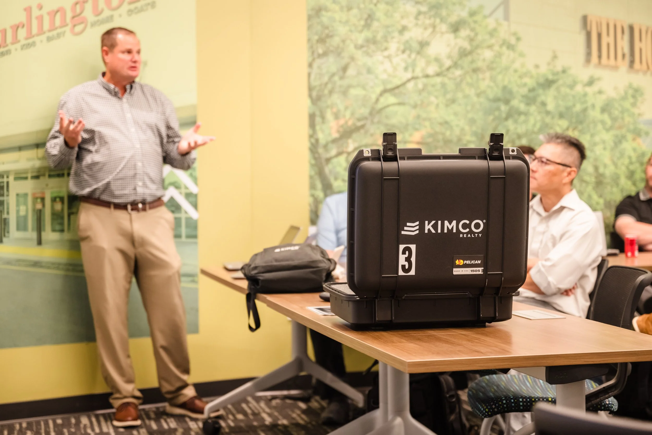 A presentation room with a man speaking to an audience, and a black Kimberley Pacific medical case on a wooden table in the foreground.