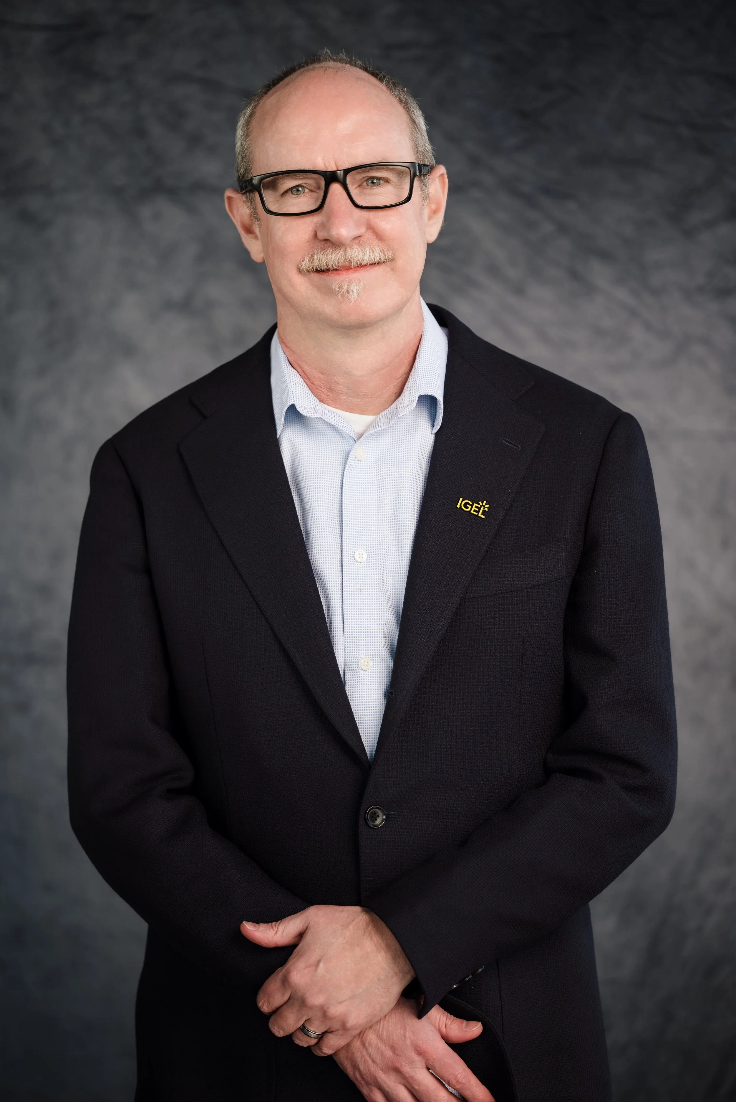 A middle-aged man with glasses, a mustache, and a goatee wearing a black blazer with an 'IGEL' pin, a light blue shirt, and standing against a gray background.