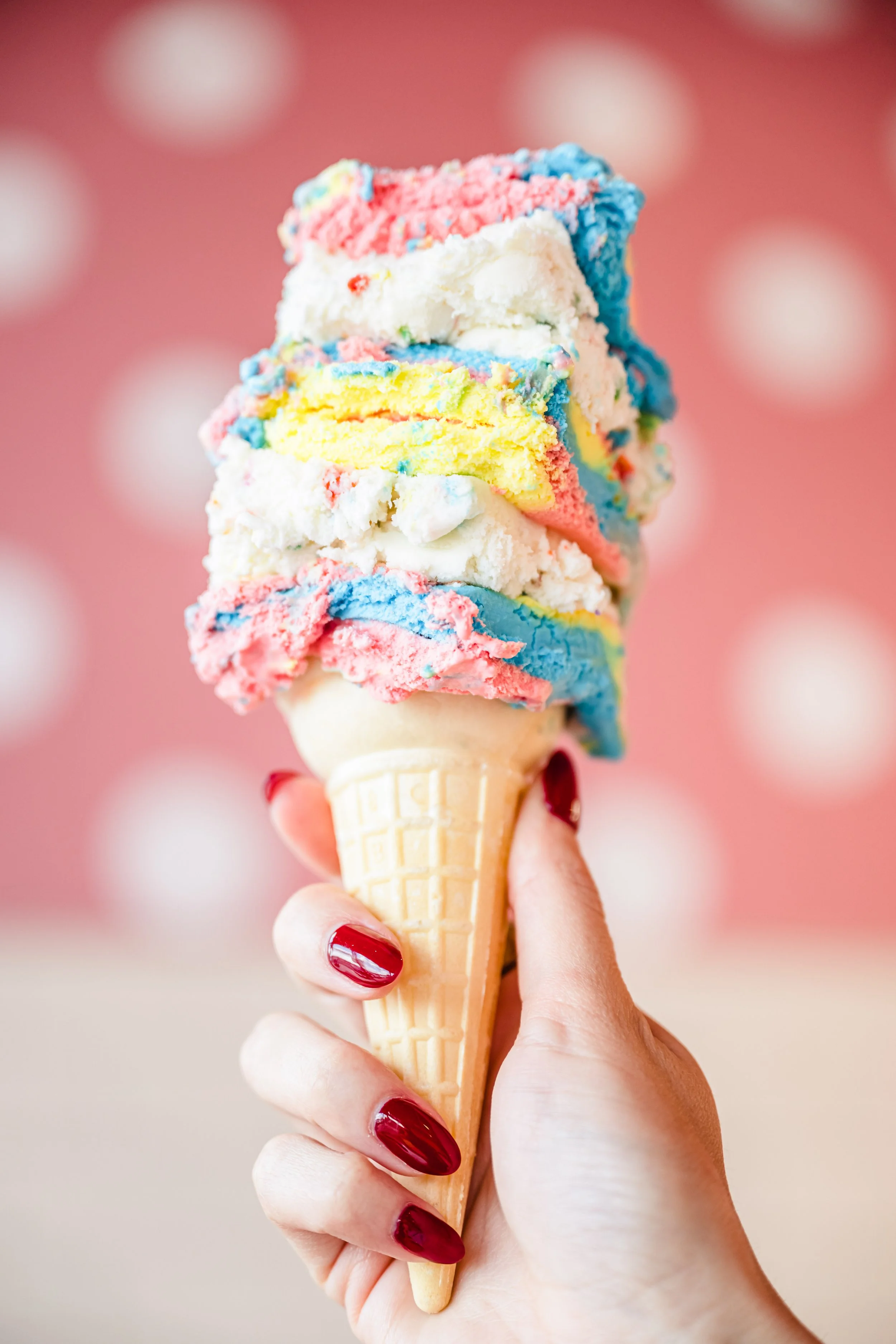 A person holding an ice cream cone with three scoops of colorful rainbow ice cream, set against a pink background.