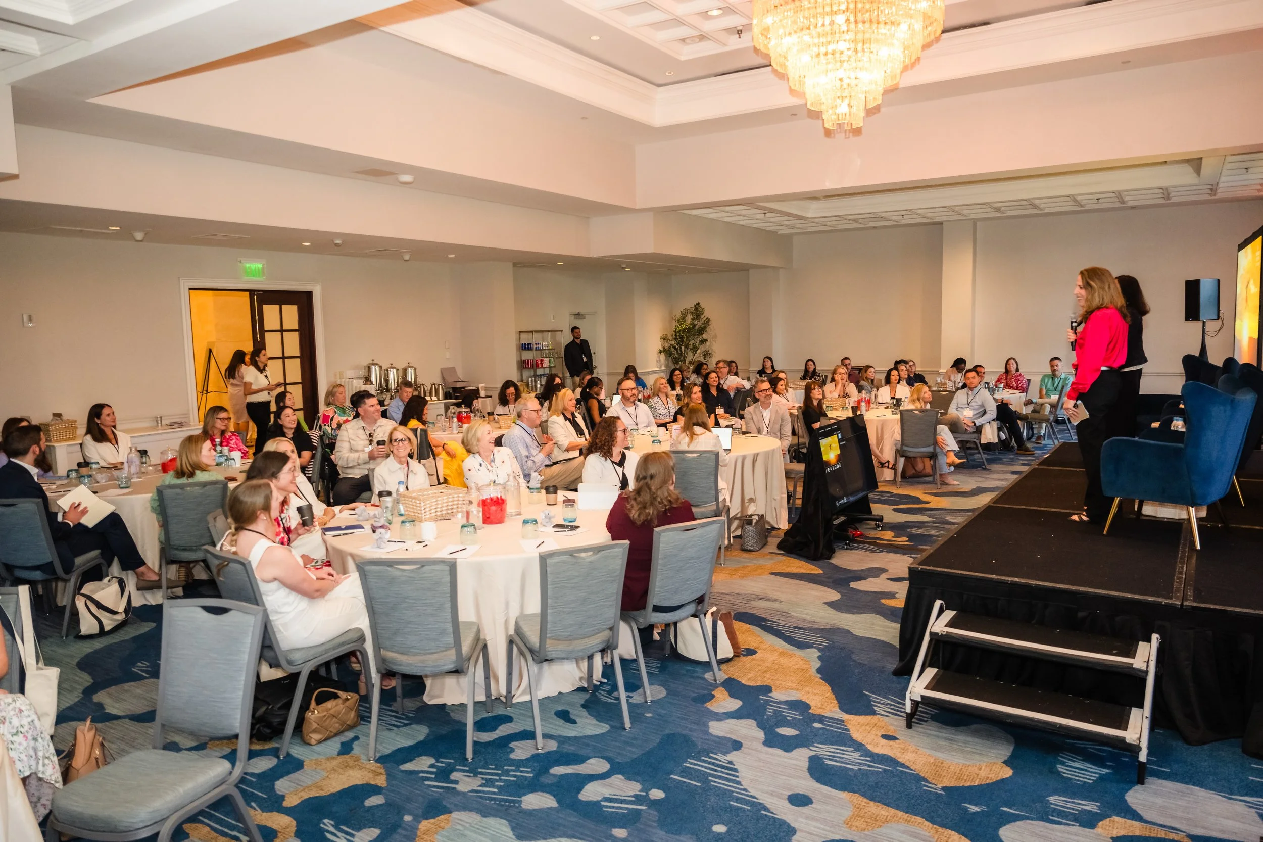A wide view of a hotel ballroom with a crystal chandelier, where dozens of attendees sit at round tables watching a speaker in a coral blouse present from a raised stage, with a presentation screen and blue velvet chairs visible on stage right.