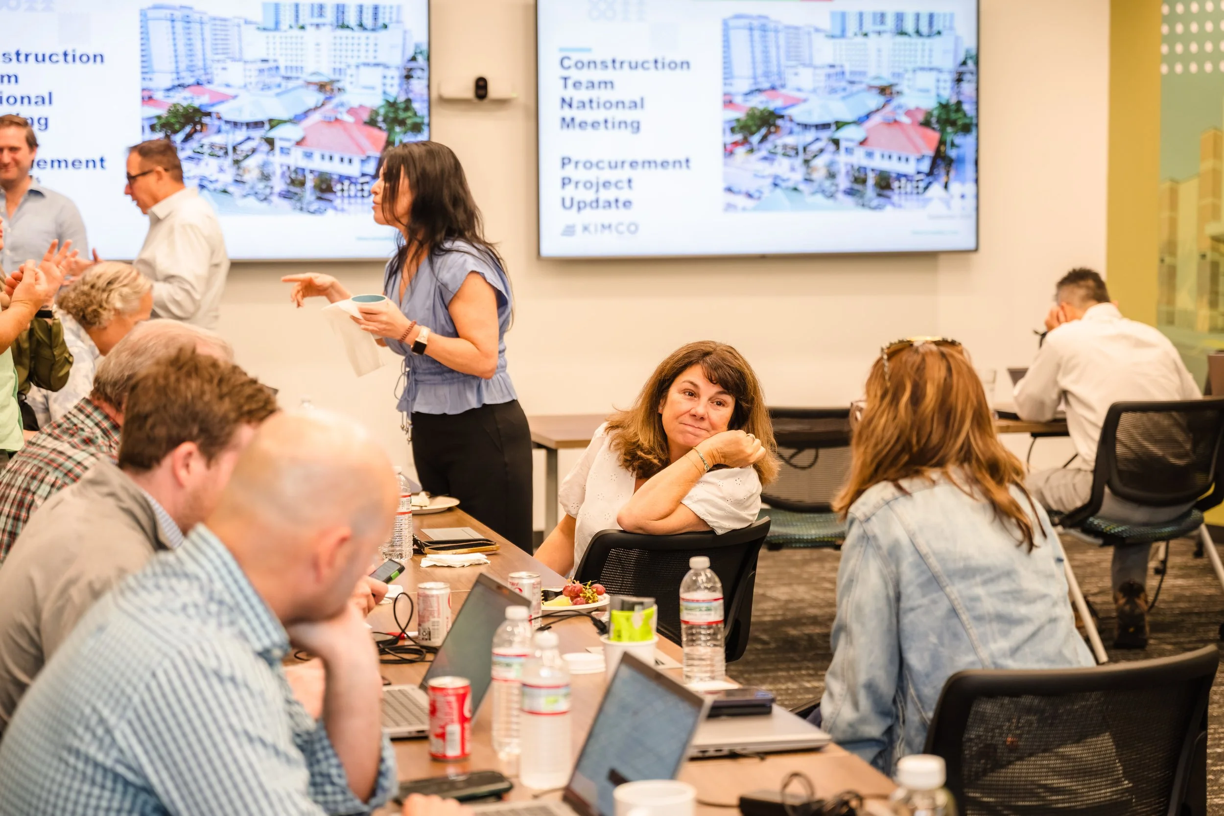 A group of people attending a construction team meeting in a conference room, with some listening, working on laptops, and one woman standing near the screen.