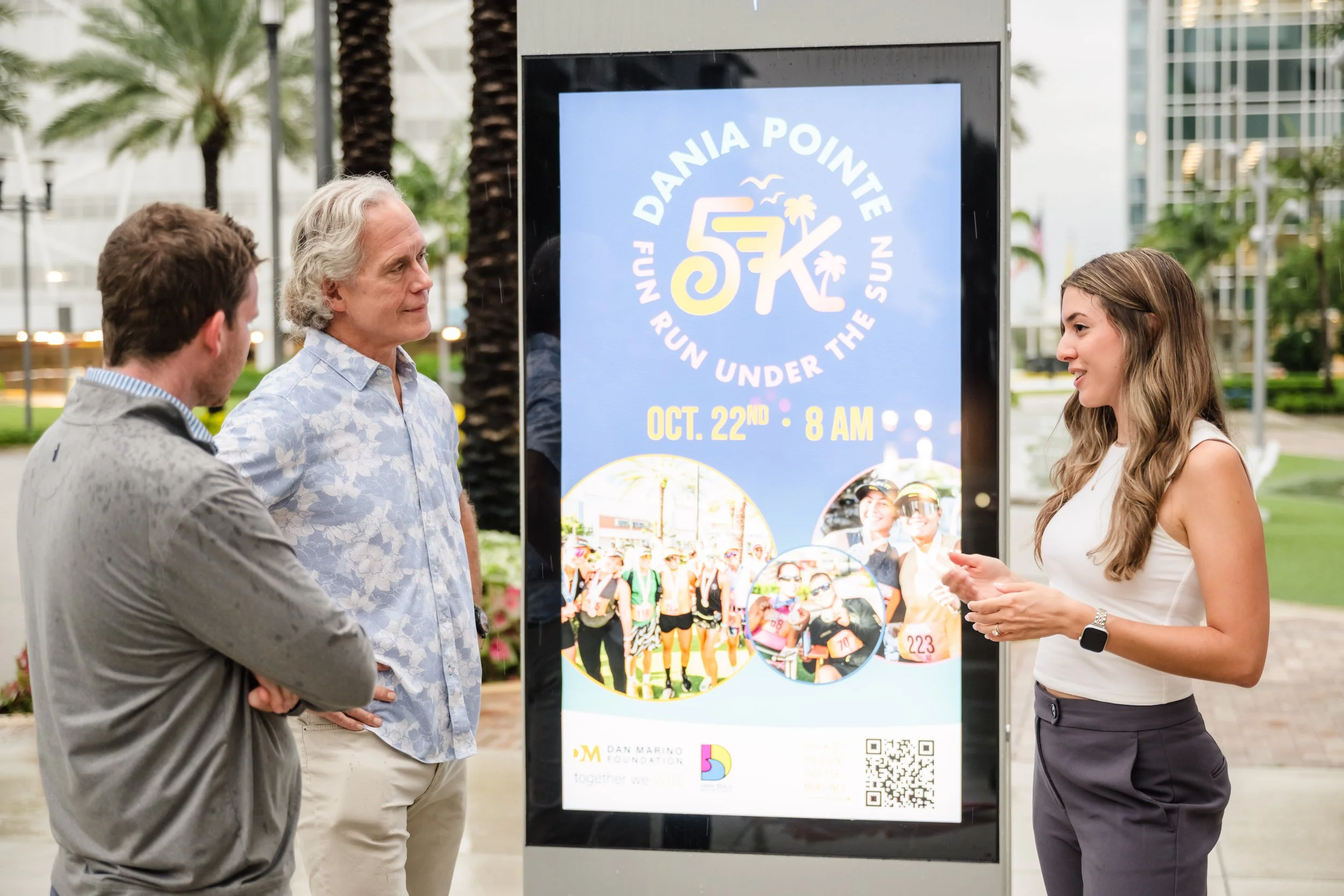 Three people standing outdoors next to a large digital signboard promoting a running event called Dania Pointe, scheduled for October 22nd at 8 AM. The sign features images of runners and event details.
