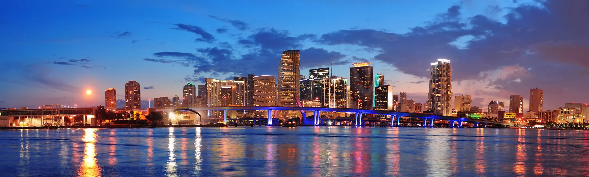 City skyline at dusk with illuminated skyscrapers reflecting in the water.
