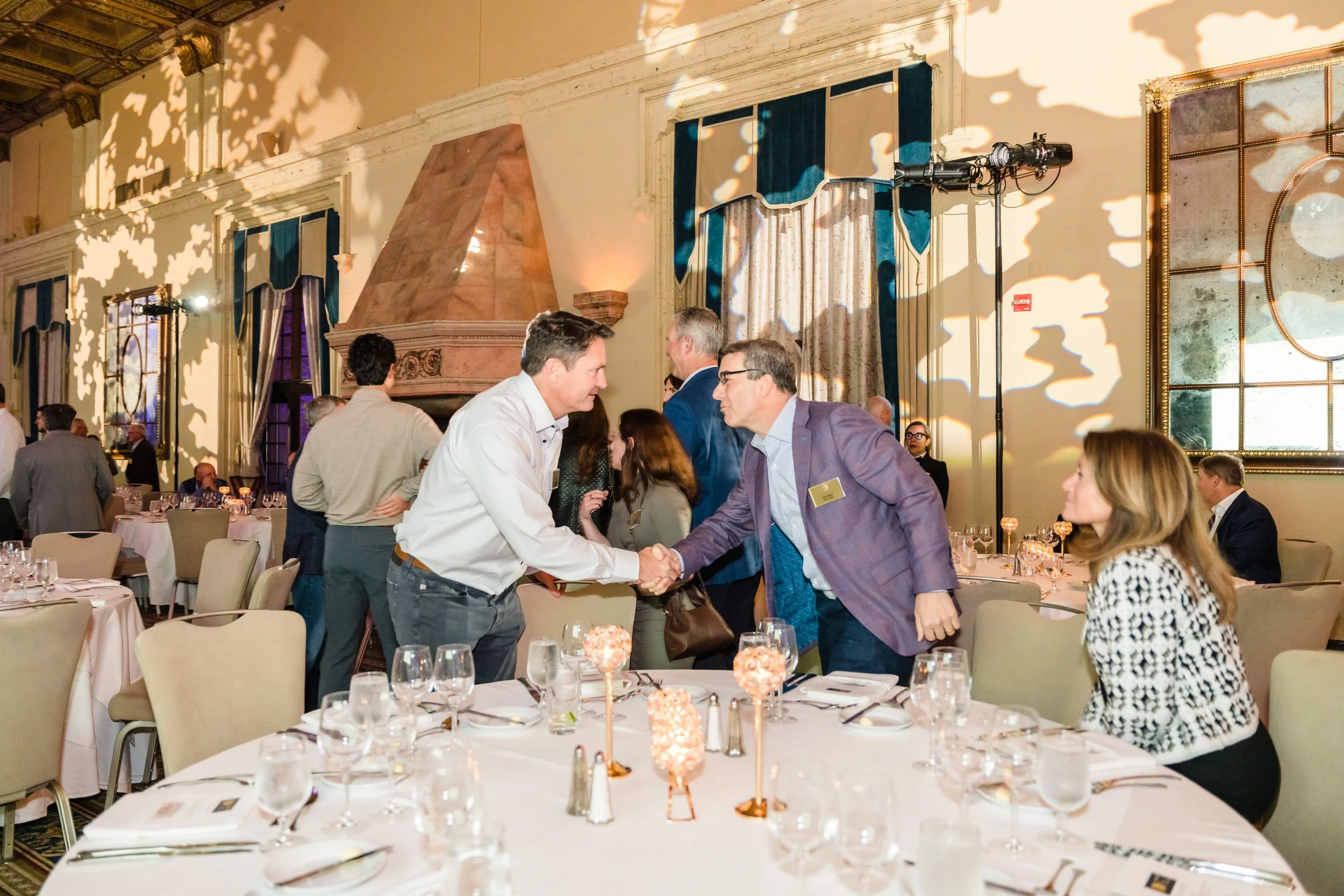 wo men shake hands between elegantly set round tables inside The Biltmore's ornate ballroom during the Mid-Market Deal Maker Symposium 2025, with other guests networking in the background amid dramatic lighting and teal drapery.