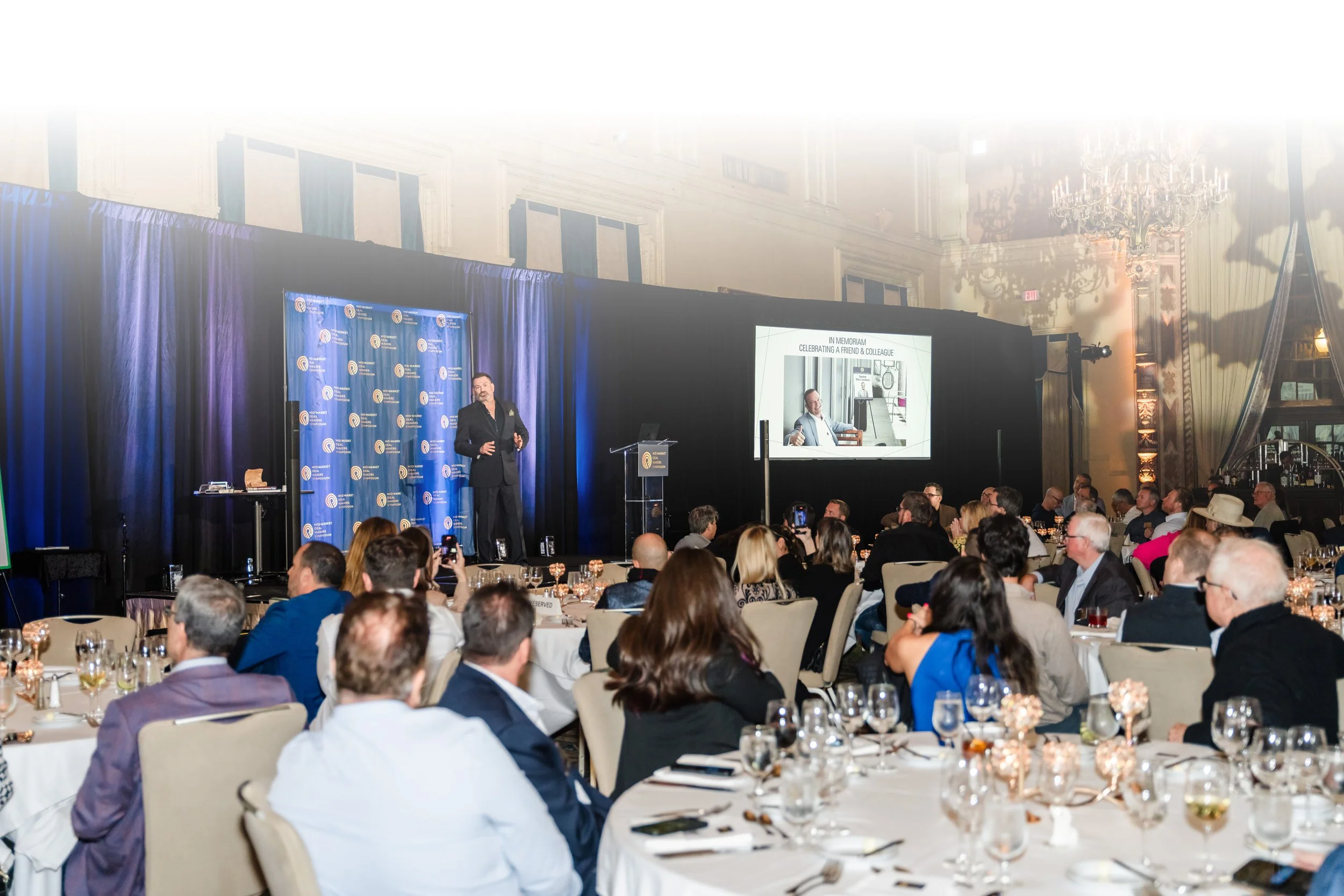 A speaker on stage at a formal event, with an audience seated at round tables in a large, ornate hall with chandeliers.
