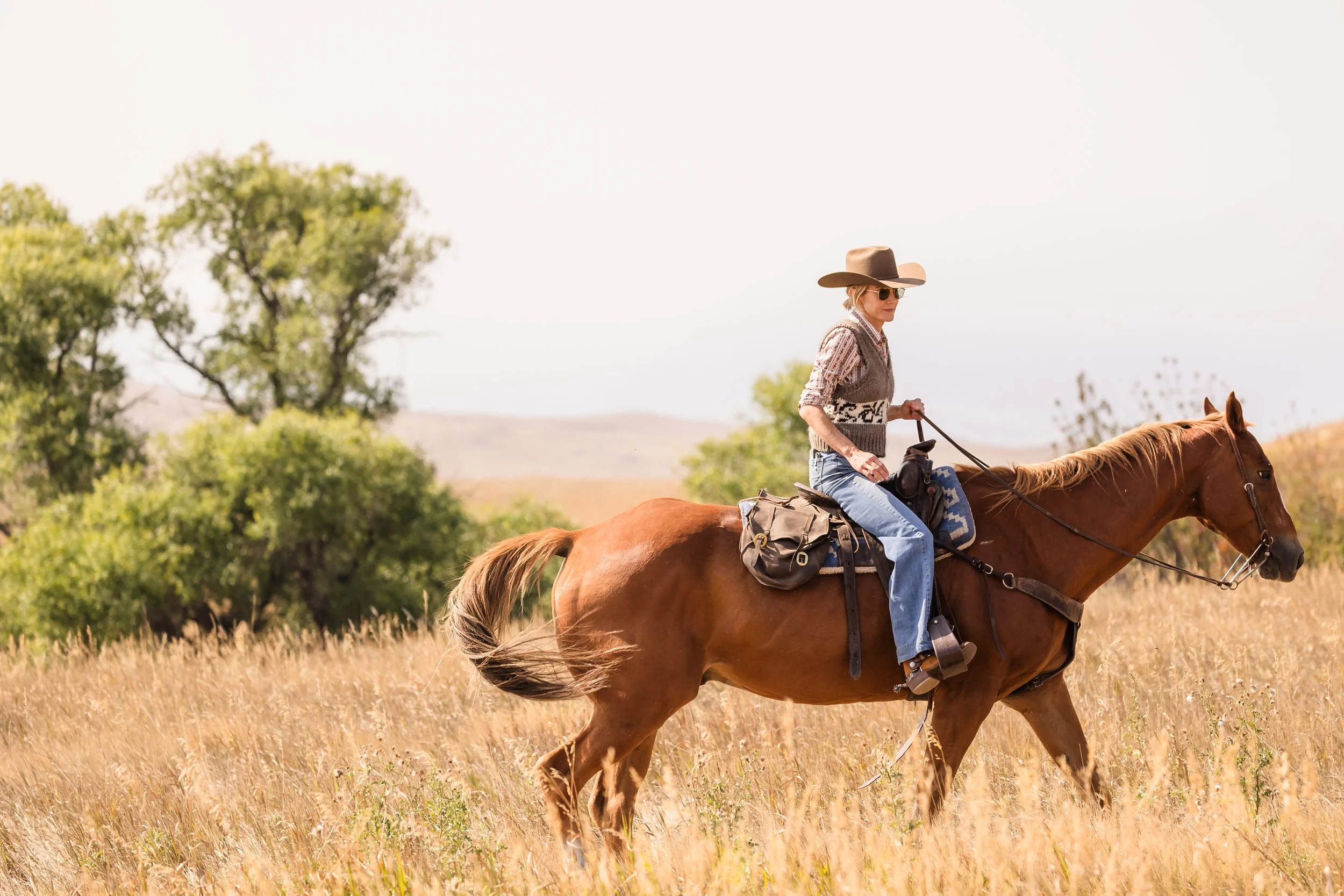 A woman riding a brown horse through a grassy field, wearing a large hat, sunglasses, a patterned sweater, and jeans, with trees and hills in the background.