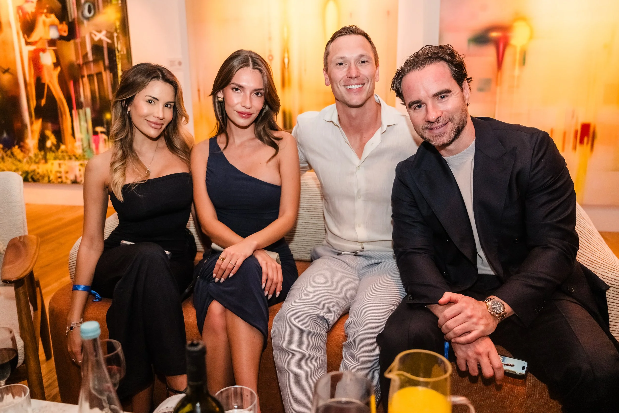 Group of four friends sitting on a sofa at a party or social gathering, smiling and posing for the photo, with drinks and bottles on the table in front of them.