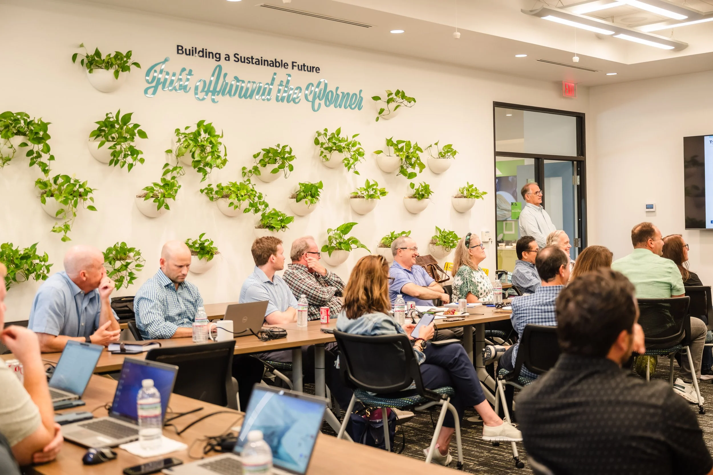 A group of people attending a presentation or meeting in a conference room with a wall decorated with potted plants and a sign that says, 'Building a Sustainable Future, Just Around the Corner.'