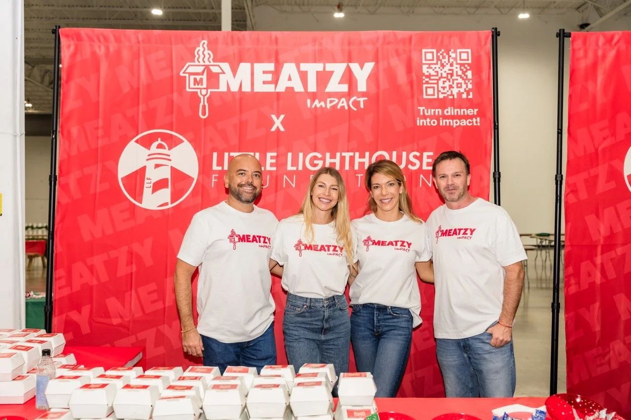 Four people wearing white T-shirts with 'Meatzy Impact' logo stand together in front of a red backdrop with a lighthouse logo and the words 'Little Lighthouse Foundation.' They are smiling, and there are boxes of food or takeout on a table in the for