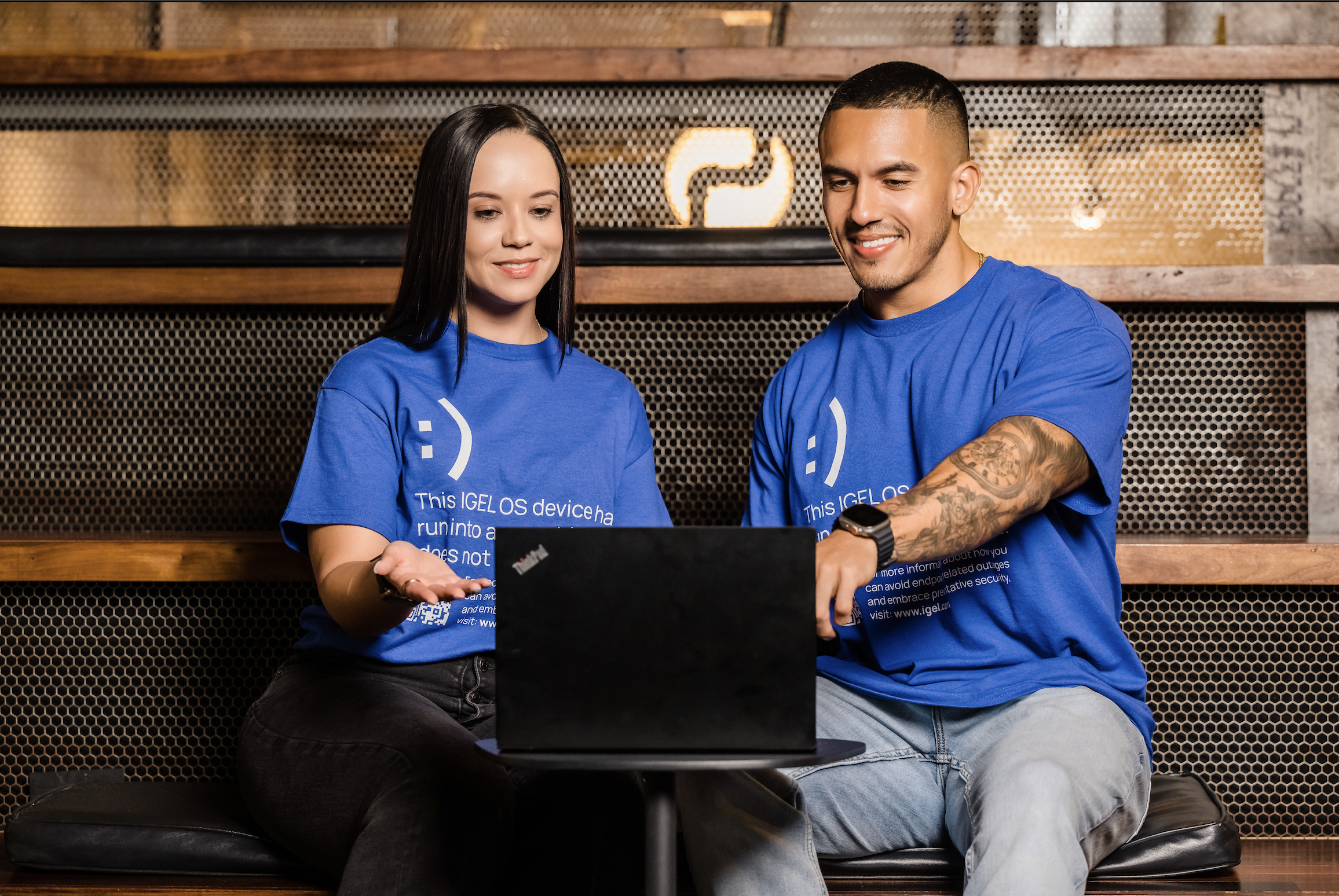 Two people sitting on a black cushioned bench in front of a wooden wall, looking at a black laptop on a small round table between them. Both are wearing blue T-shirts with white text and smiley face icons, indoors with warm lighting.