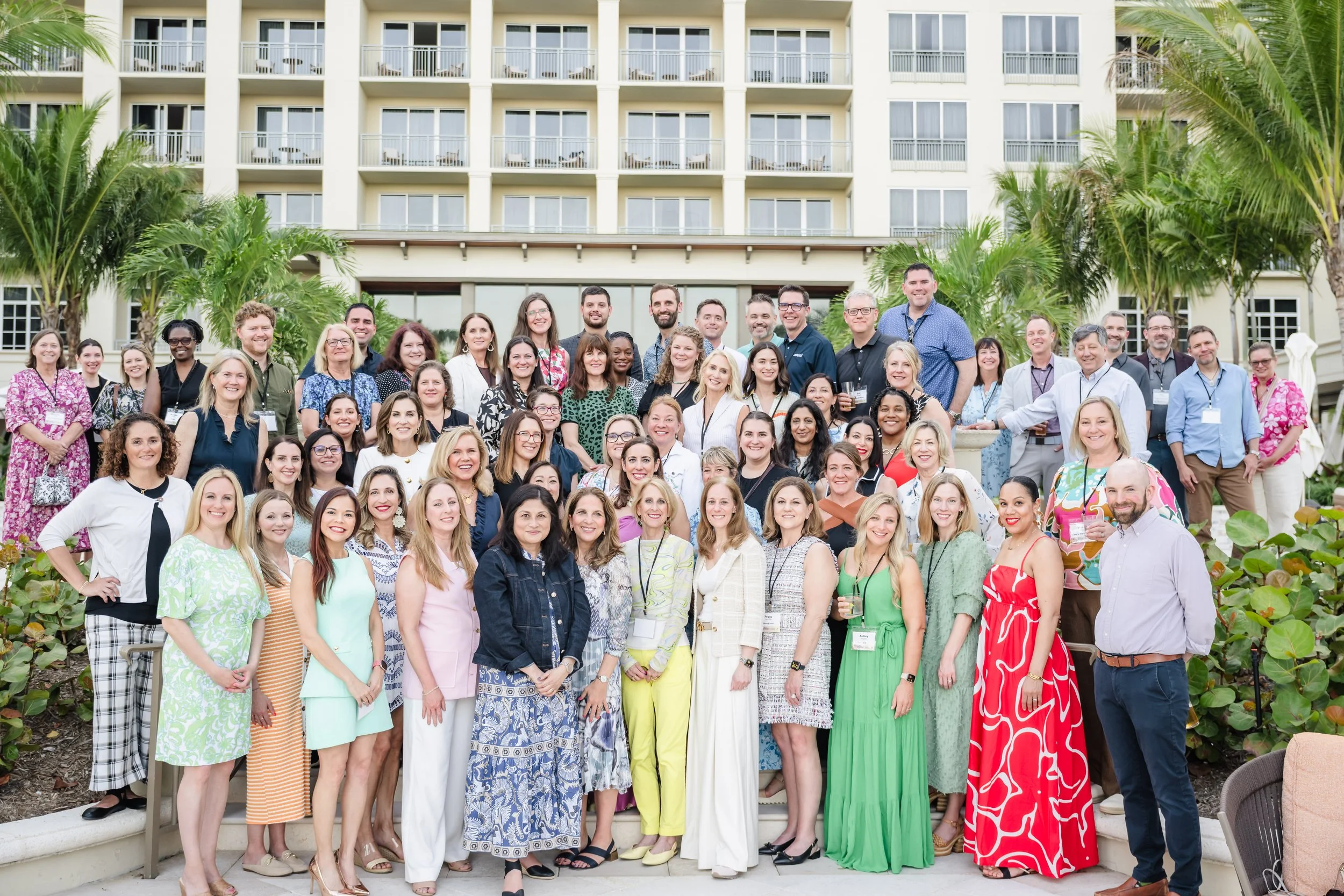 A large group of communications professionals in colorful business-casual attire stands together on outdoor steps for a group photo, with the cream-colored facade of The Ritz-Carlton, palm trees, and tropical landscaping behind them.