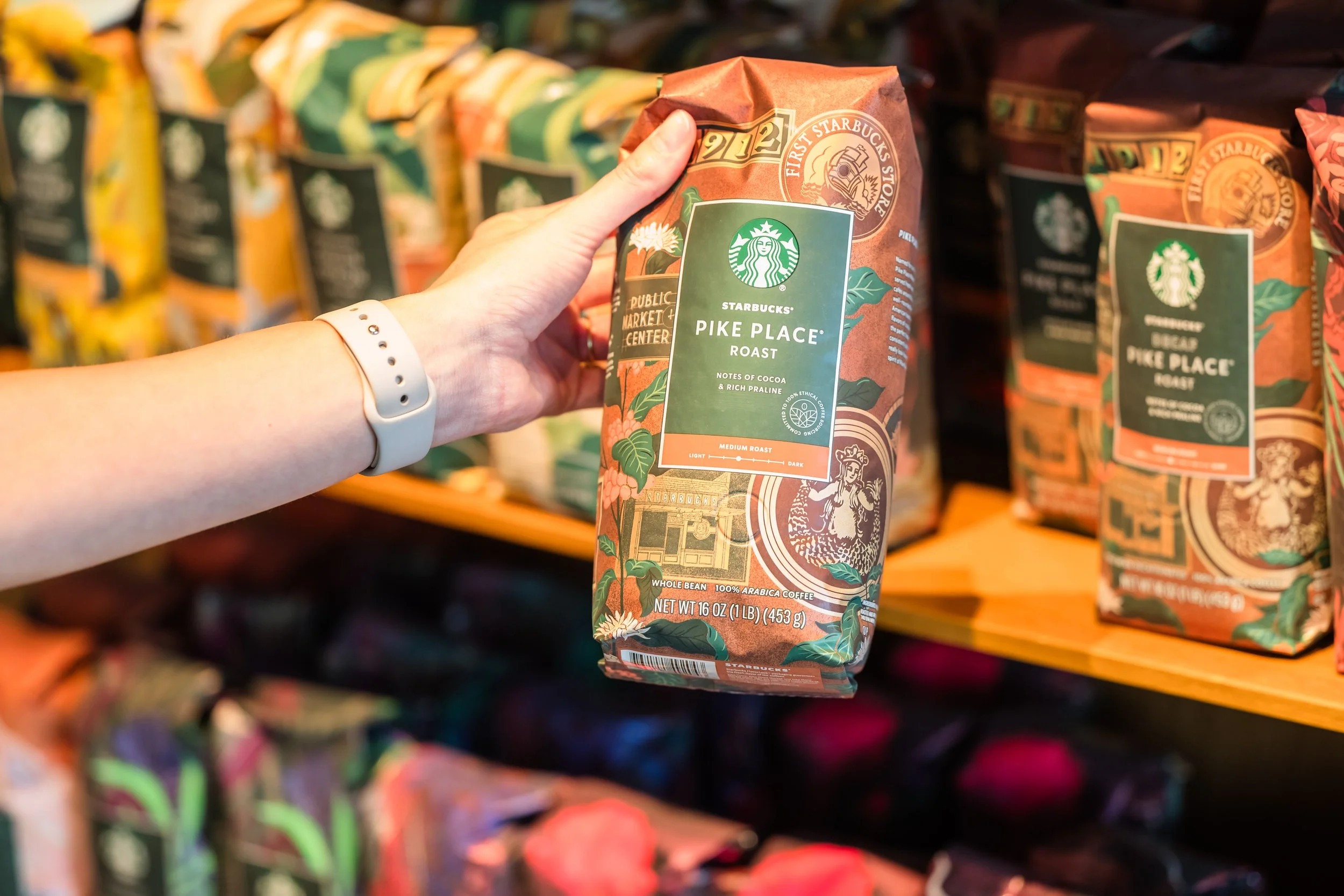 A hand holding a bag of Starbucks Pike Place Roast coffee in a grocery store aisle.