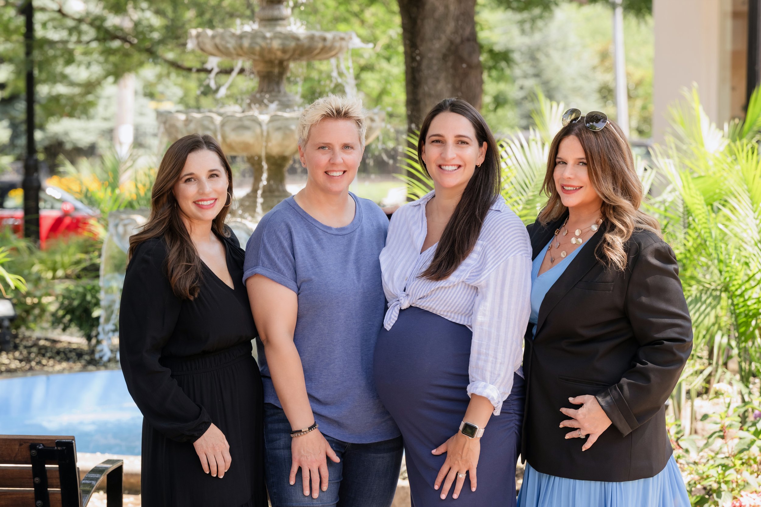 Four women standing together outdoors in front of a fountain, smiling, with greenery behind them.