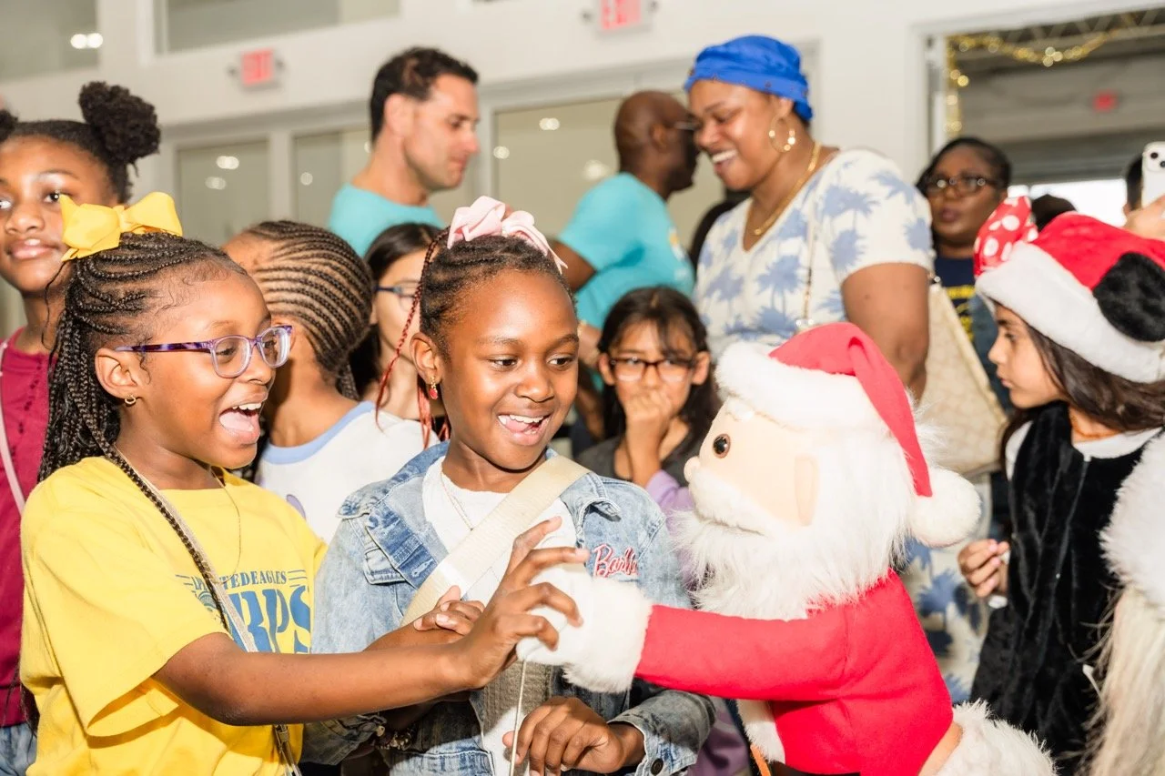 Children and adults gathered around a doll in a Santa costume at a holiday event, smiling and interacting with the Santa figure.