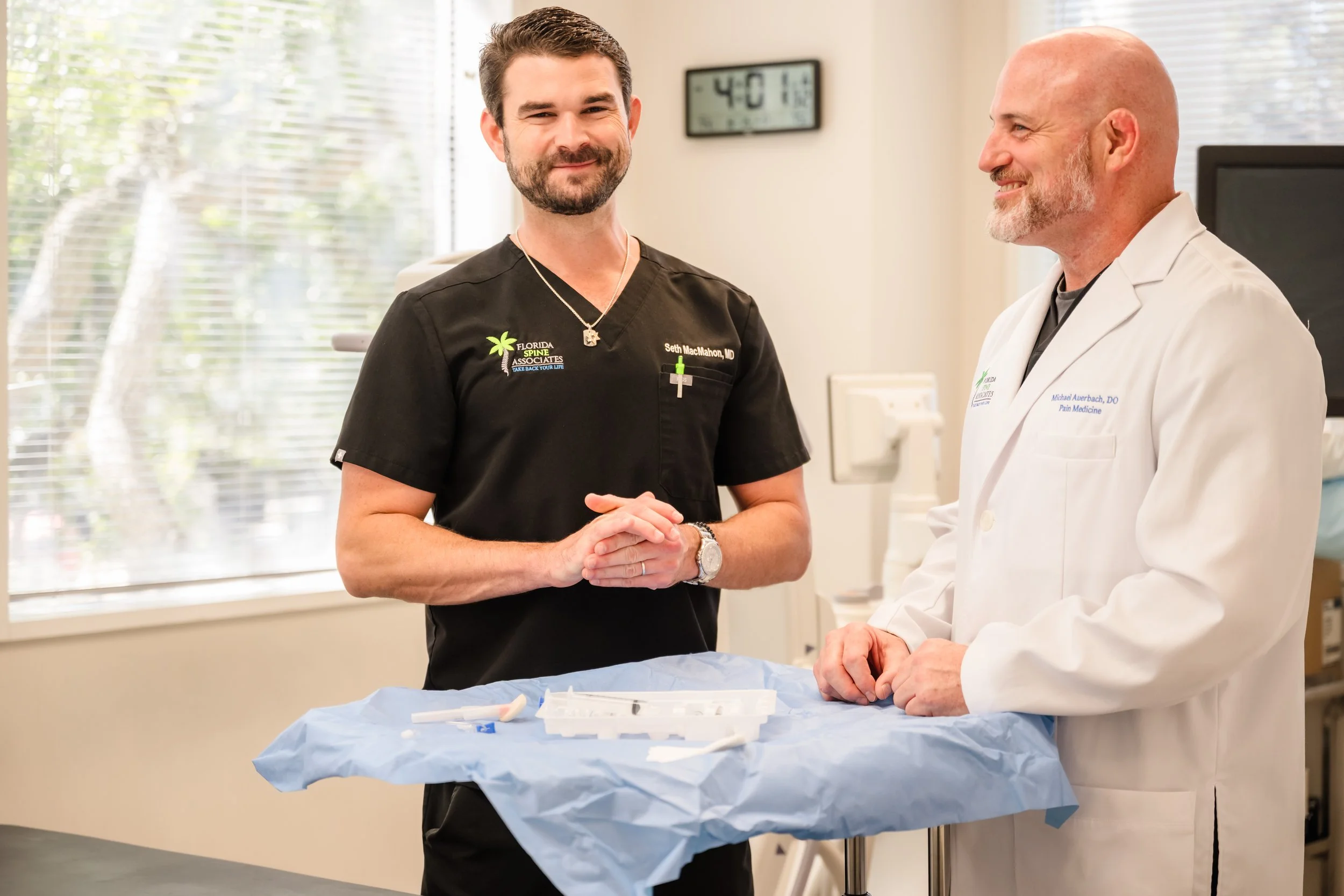 Two doctors, one in black scrubs and the other in a white coat, standing in a medical office with a tray of medical supplies on a blue cover, smiling and talking.