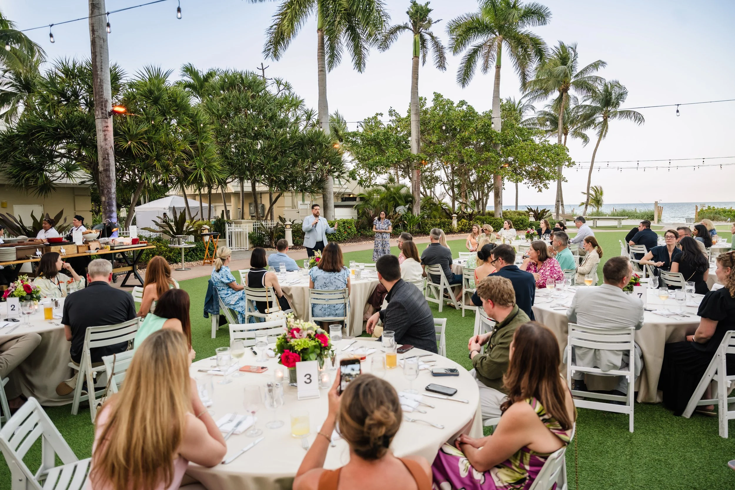 Attendees seated at numbered round tables with white linens and pink floral centerpieces on a manicured lawn listen to two speakers with microphones, framed by palm trees, string lights, and an ocean view at dusk.