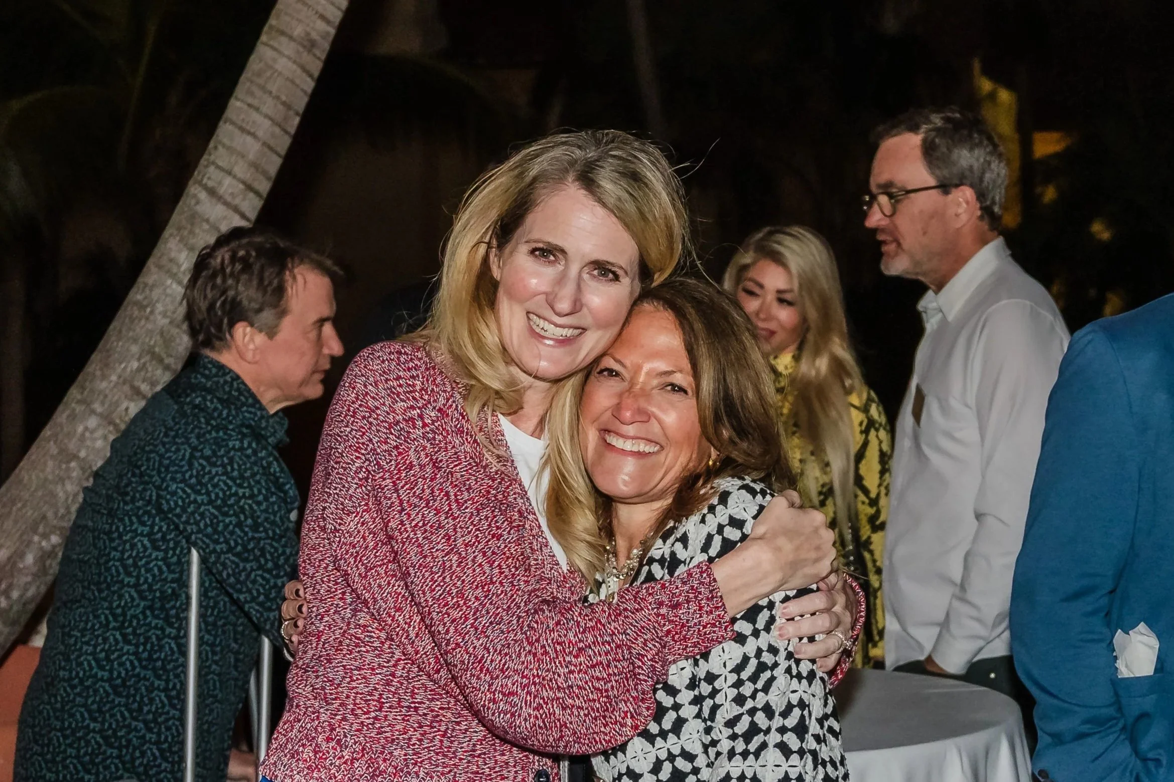 Two women hug and smile for the camera on the patio during an outdoor nighttime reception at The Biltmore, with palm trees, cocktail tables, and other guests mingling in the background.