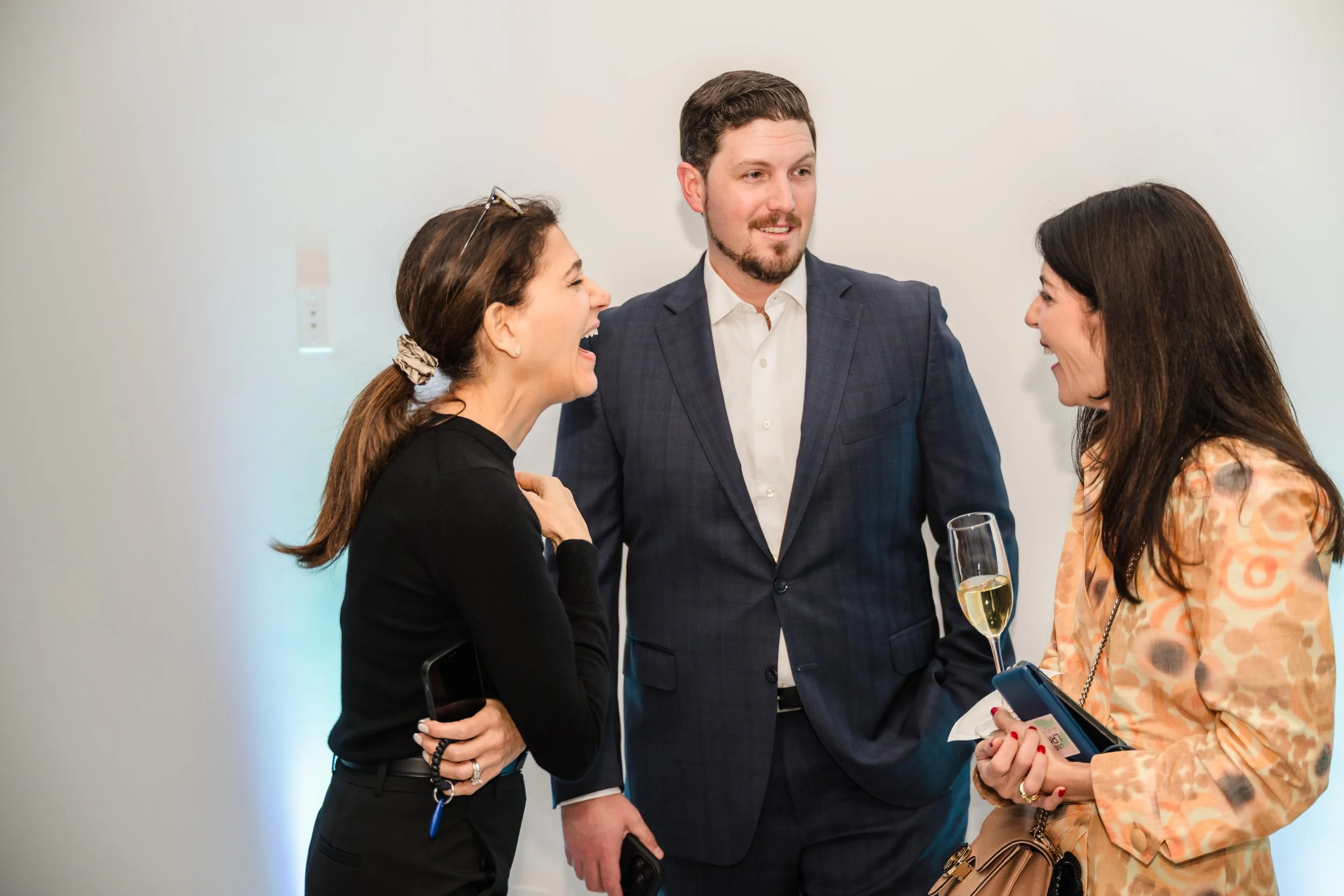 Three people talking and laughing at a social event, with one woman holding a glass of champagne
