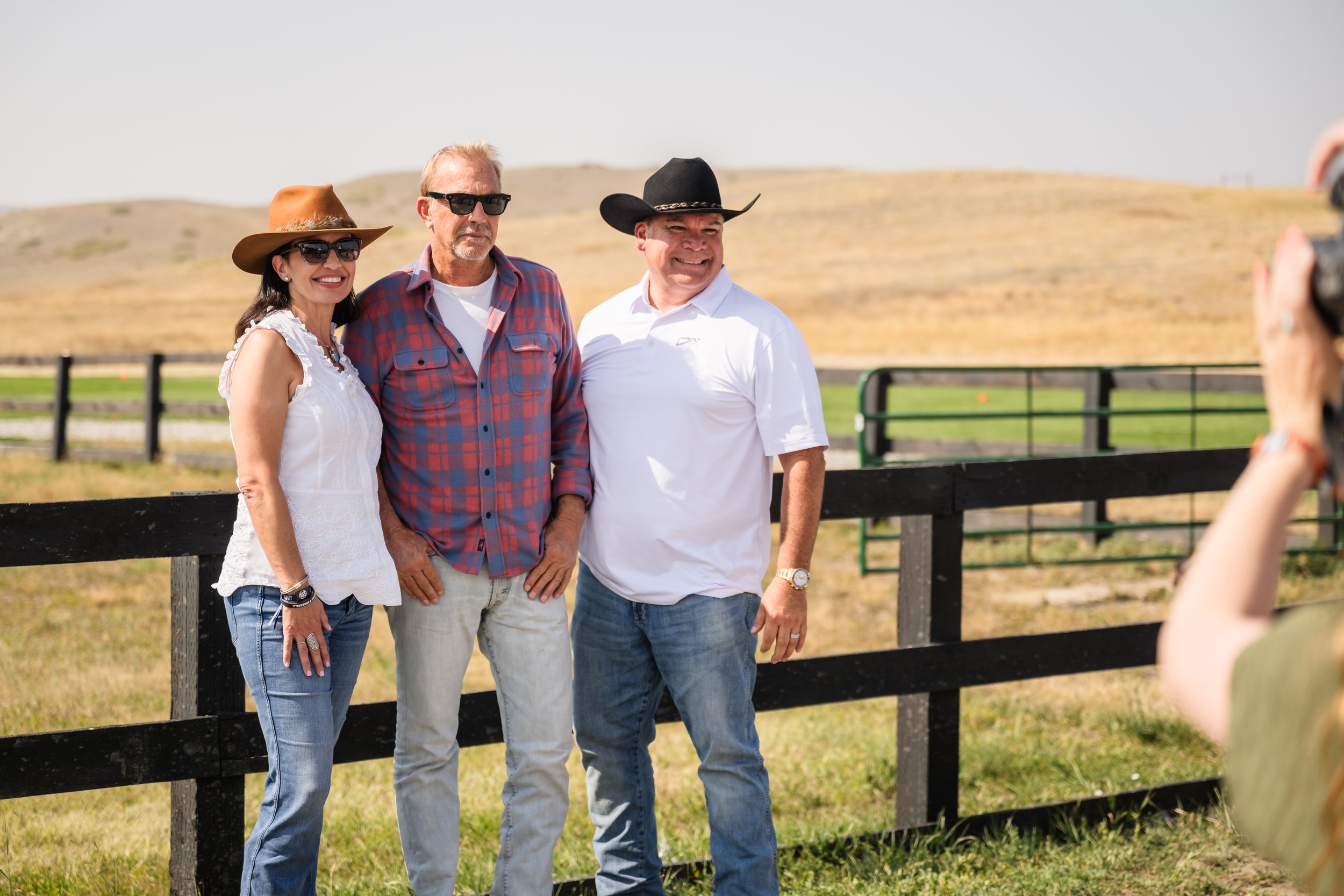 Three people taking a photo outdoors in a rural area with hills in the background. One woman and two men, all wearing casual shirts and hats, smiling.
