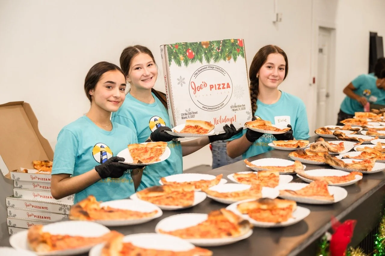 Three young women in light blue shirts and black gloves holding plates of pizza, standing behind a table filled with slices of pizza, with a sign for Joe's Pizza, at a holiday event.