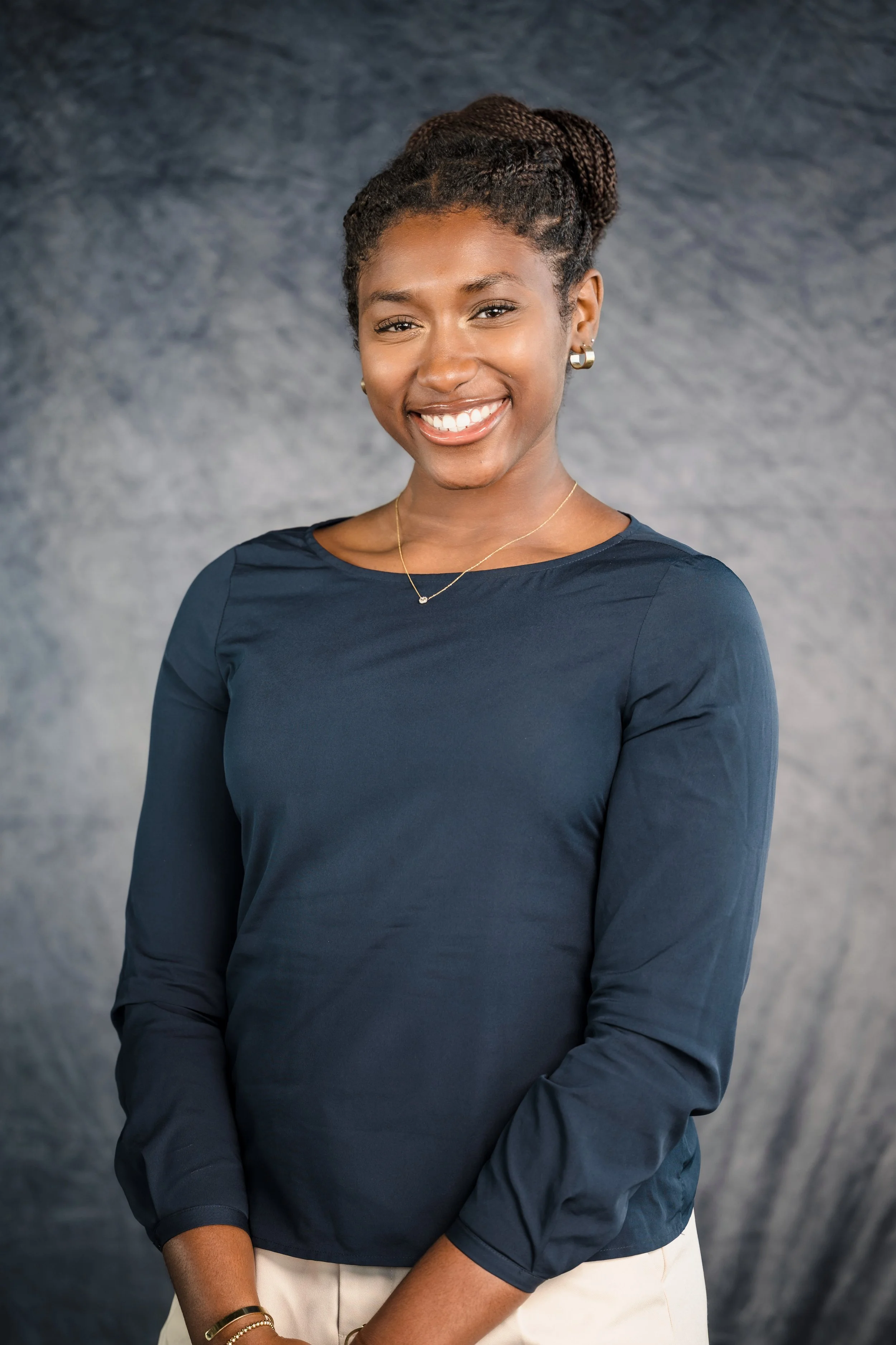 Young African American woman smiling, wearing a dark long-sleeve shirt and jewelry, against a gray textured background.