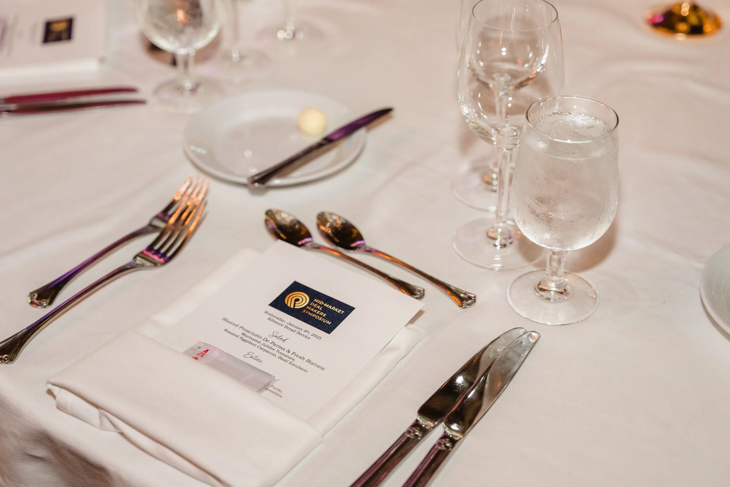 Close-up of a formal place setting featuring a white linen napkin topped with a printed dinner menu bearing the Mid-Market Deal Makers Symposium logo, surrounded by polished silverware, crystal glassware, and water glasses on a white tablecloth.