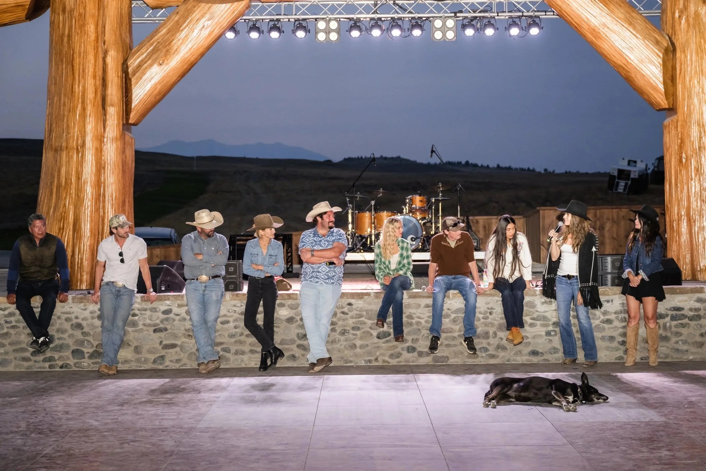Yellowstone TV cast members sitting and standing along the edge of an outdoor concert stage in western wear at dusk with Montana hills.