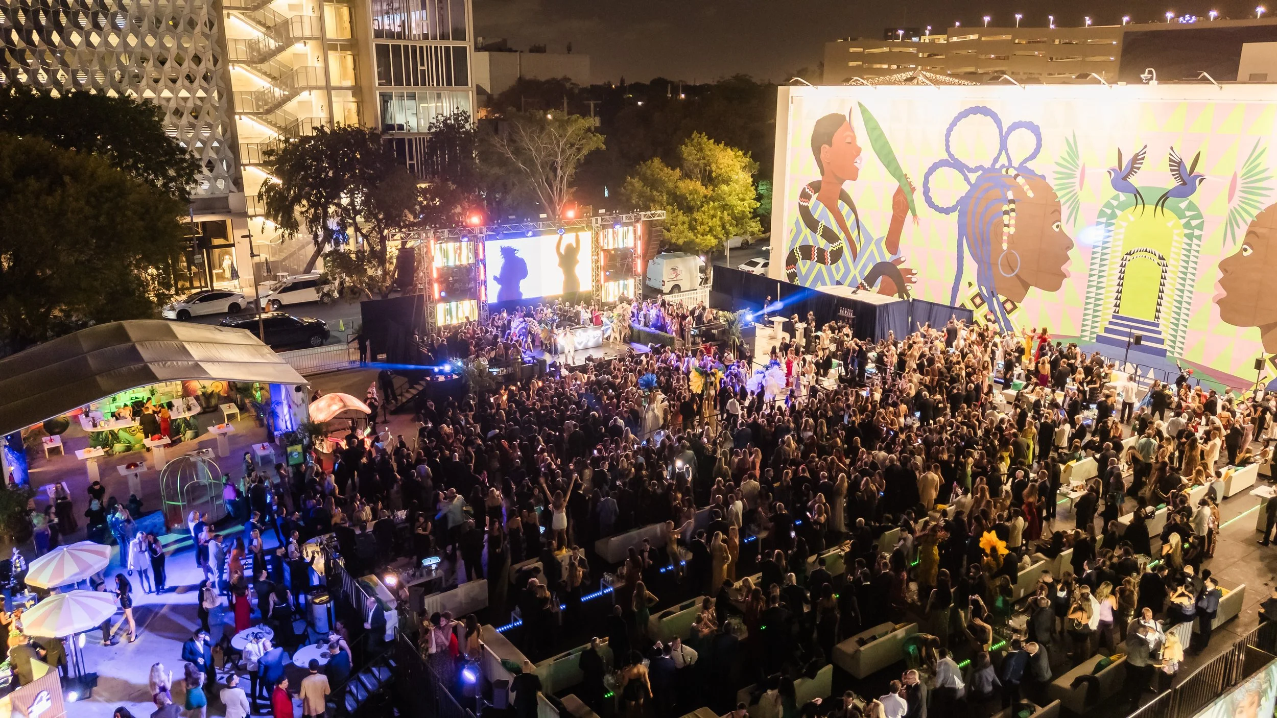Aerial nighttime view of a large crowd gathered at Jungle Plaza in the Miami Design District during the Little Lighthouse Foundation 2025 Hearts and Stars Gala with a lit stage and colorful mural in the background