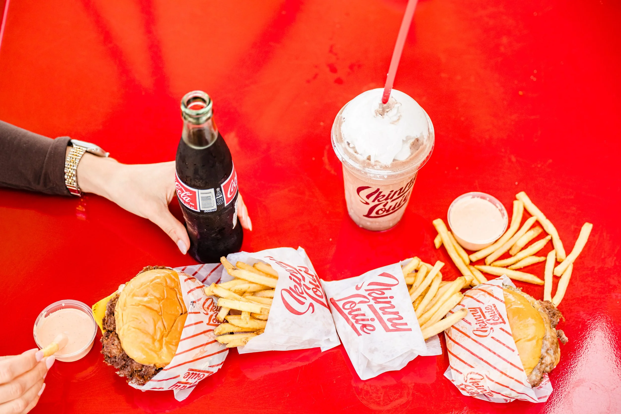 Two cheeseburgers, plates of French fries, dipping sauces, a Coke bottle, and a milkshake on a red table