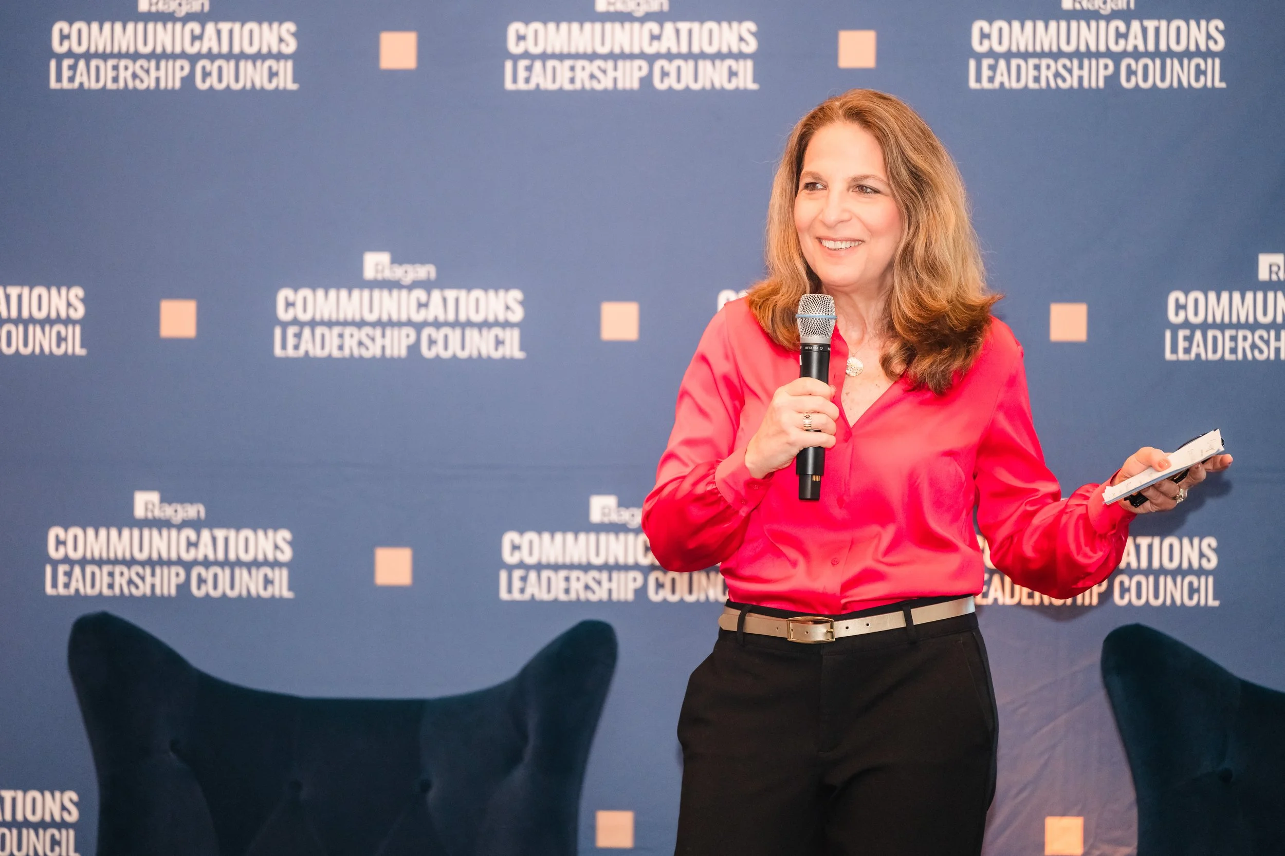 A woman in a coral blouse holds a microphone and note cards while speaking on stage in front of a blue Ragan Communications Leadership Council branded backdrop, with dark lounge chairs visible behind her.