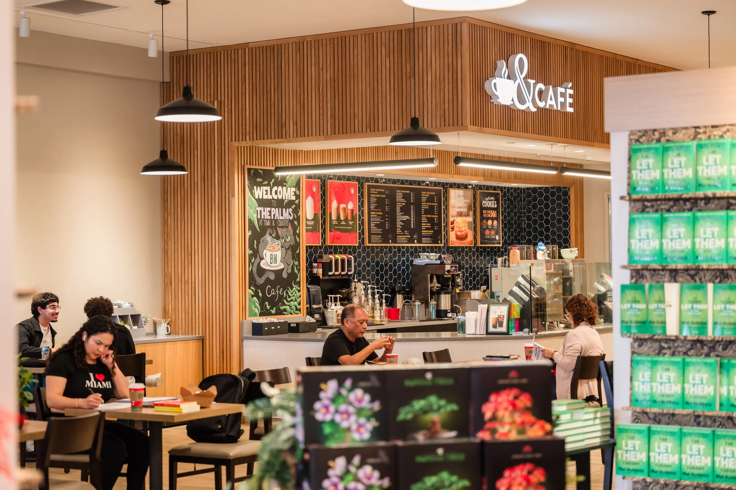 Inside a coffee shop with customers sitting at tables. The counter has a menu above it, and a chalkboard sign says 'Welcome to the Palms at Town & Country.' There are shelves with books or products in the foreground.