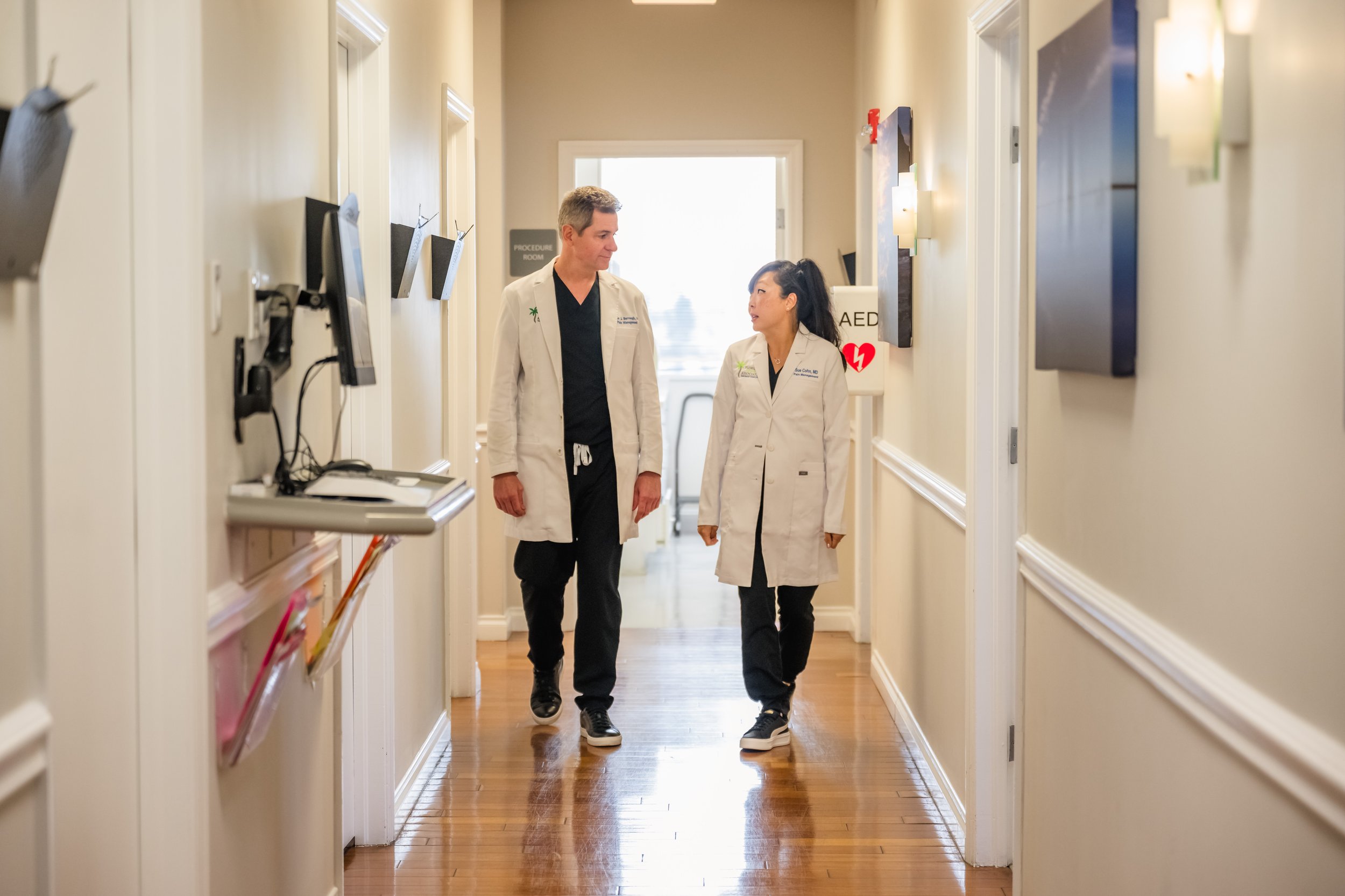 Two healthcare professionals, a man and a woman, walking down a hospital corridor, engaged in conversation. Both are wearing white lab coats and black scrubs, with the woman on the right also wearing sneakers. The corridor has wall-mounted monitors a