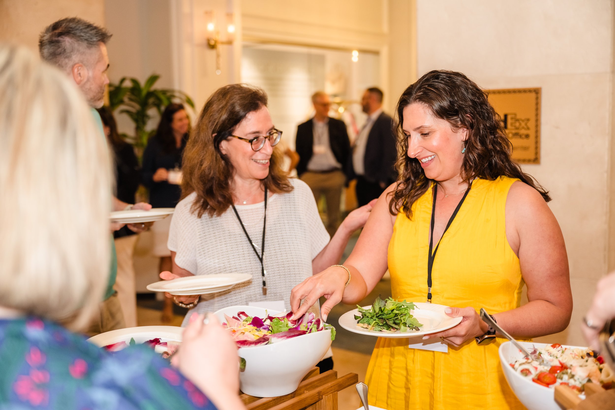 Two smiling women wearing conference lanyards serve themselves from a colorful salad buffet while chatting with fellow attendees in an elegant hotel setting with warm lighting and tropical plants in the background.